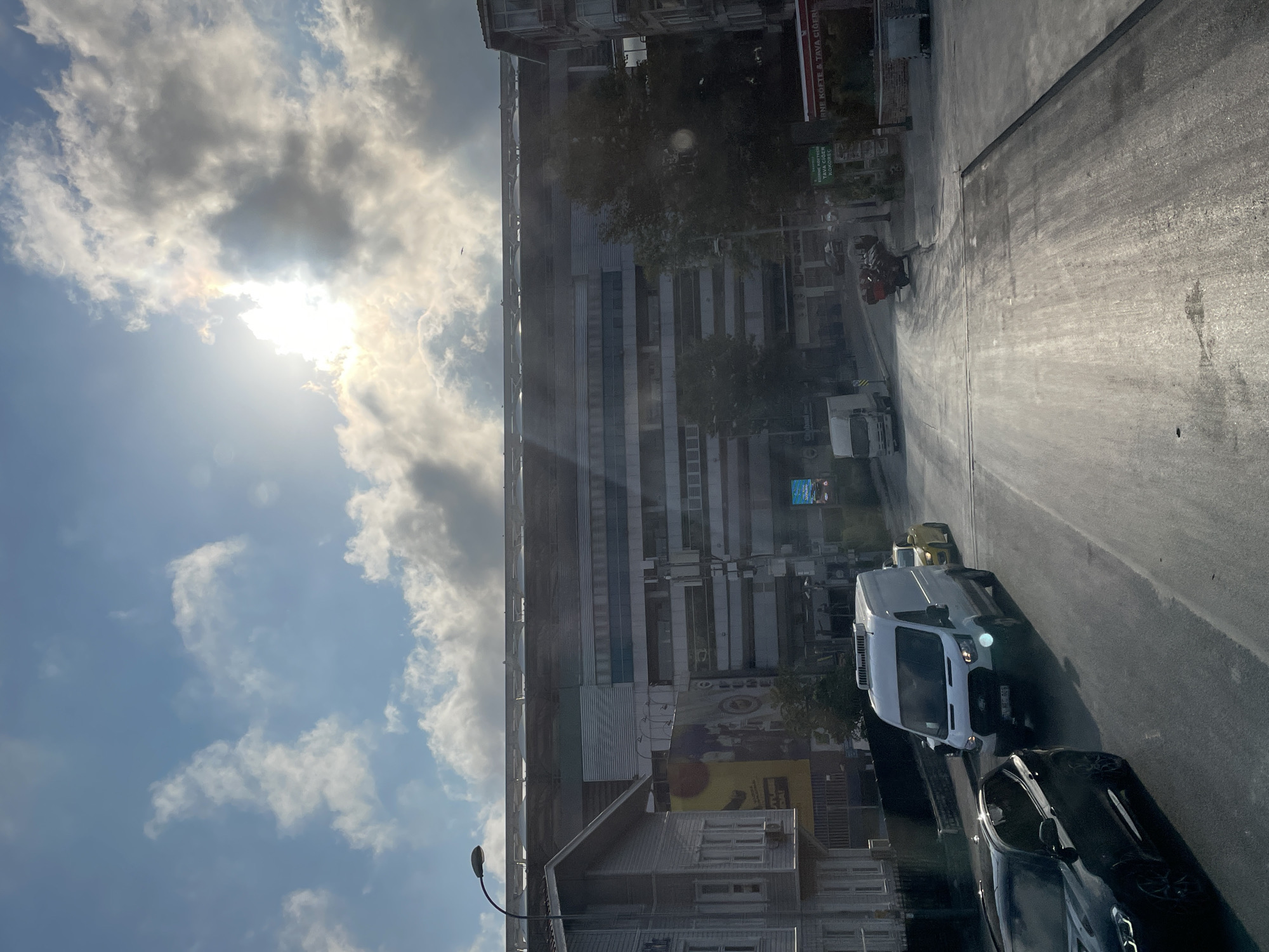 In Kadıköy, Turkey, a street scene unfolds under a bright, partly cloudy sky. The sun is partially obscured by clouds, casting a bright glow and lens flare. The street is paved with asphalt and appears wet or recently washed, reflecting the sunlight. A black SUV is in the foreground on the left, followed by a white van. Behind the van, a yellow car is visible, and further down the street, there is a motorcycle carrying a load. In the background, a large, multi-story building, likely a stadium or arena, dominates the skyline with its modern architectural style. To the left of this structure is a smaller, traditional-looking house. Signs are visible on the street, including one for "KÖFTE & TAVA CIGER." The overall impression is a daytime urban environment with vehicles on the road and a prominent building in the background.