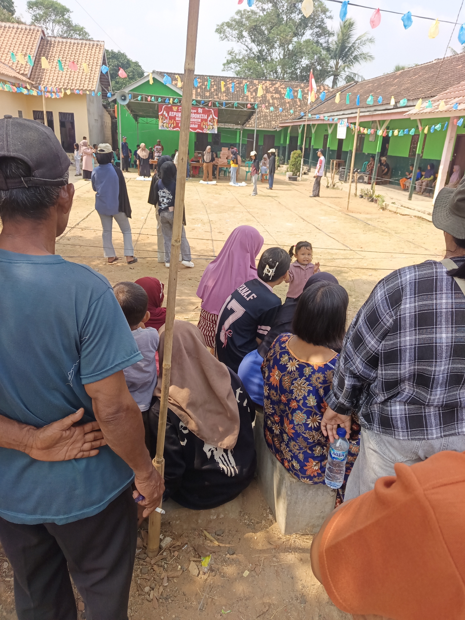 The image depicts an outdoor gathering in what appears to be a village or community area in Terbanggi Marga, Indonesia. In the foreground, several people are seen from the back, including a man wearing a blue t-shirt and a man in a plaid shirt. They are observing an event taking place in a central courtyard.
The courtyard is filled with people, some standing and others sitting on a raised concrete structure. A stage or platform is set up at one end, decorated with a banner and colorful flags. People are gathered around the stage, suggesting an event such as a performance, speech, or competition.
Buildings with tiled roofs are visible in the background, characteristic of Indonesian architecture. The ground is a dirt or sand surface, common in outdoor community spaces. The atmosphere appears festive, with decorations suggesting a celebration or community event. The time of day seems to be daytime, with bright sunlight casting shadows.