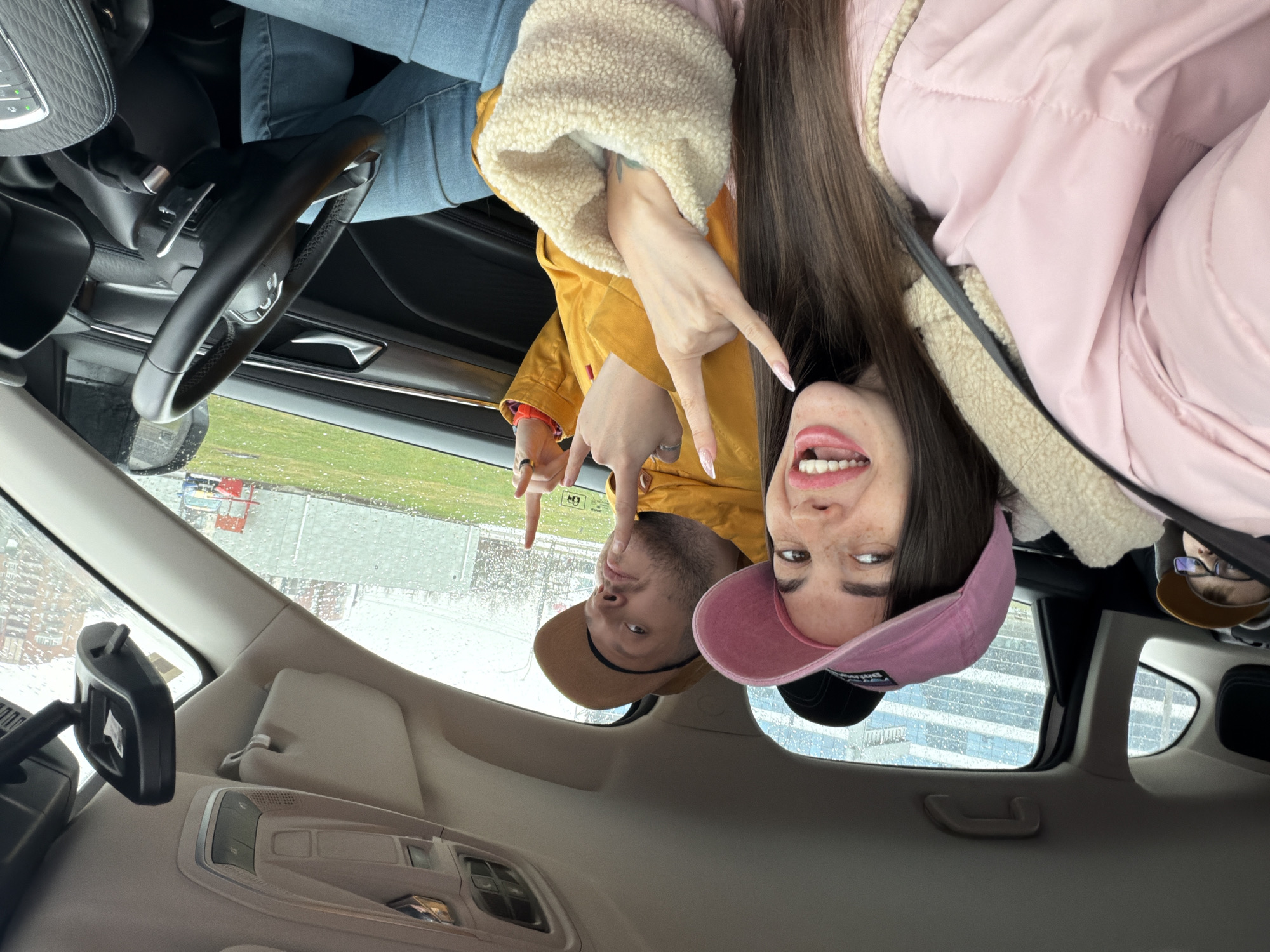 Two individuals, a male and a female, are visible inside a car. The female is wearing a pink hat and a pink coat with a faux fur collar. The male is wearing a tan hat and a yellow jacket with a faux fur collar. Both are making hand gestures. The location appears to be Saint Petersburg, Russia, as evidenced by the visible architecture and weather conditions outside the car window. It seems to be raining. The interior of the car is visible, including the steering wheel, rearview mirror, and dashboard.