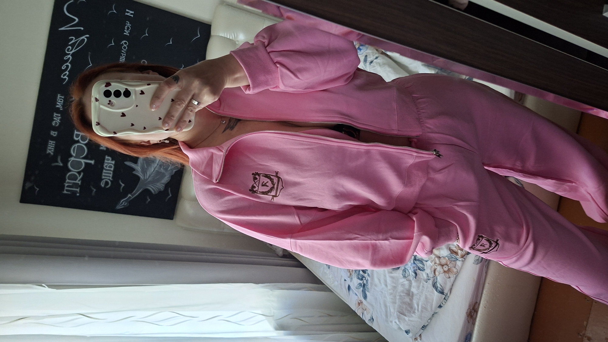 A woman with red hair is taking a selfie in a bedroom. She is wearing a pink tracksuit with a shield logo on the jacket and pants. The tracksuit is zipped down to reveal a black bra. The room appears to be decorated with a chalk board behind the woman. The woman is standing in front of a bed, which is covered in a floral print comforter. She is holding a phone in her right hand, covering her face. The image is taken from a slightly low angle, looking up at the woman. The woman has tattoos on her arm and neck. She is wearing a silver ring on her left ring finger and dangling earrings. The overall lighting suggests it is daytime, with natural light coming from a window on the left side of the image. The background is slightly blurred, indicating a shallow depth of field. There is a mirror on the right side of the image, reflecting part of the room and the woman. The location is identified as Myadzyel, Belarus.