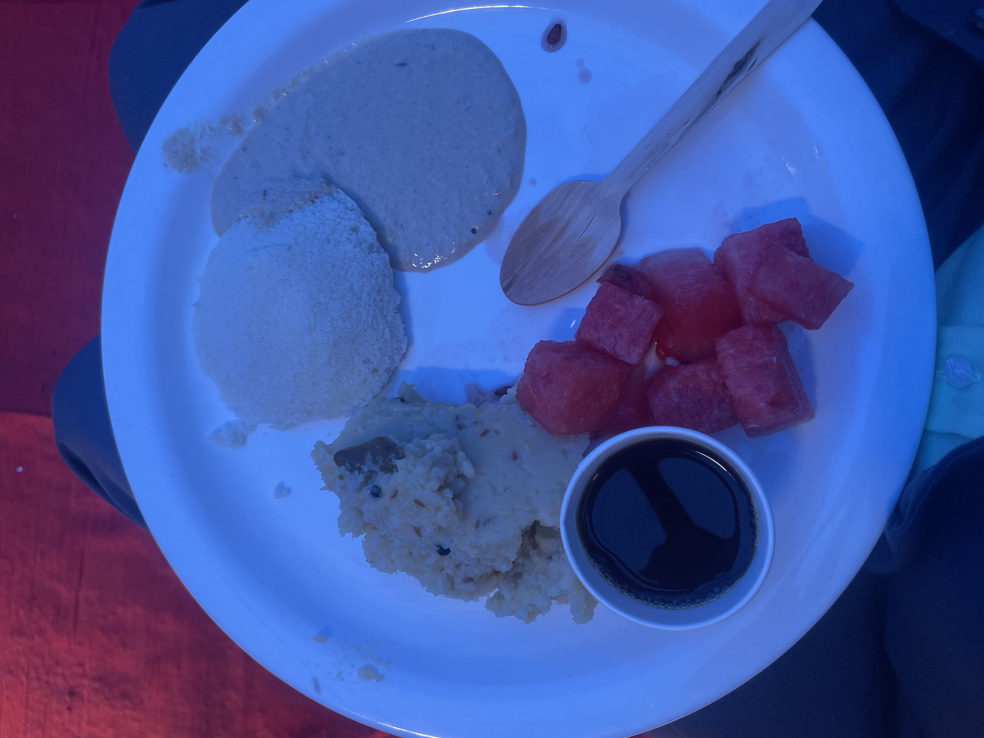 The image shows a white plate with several food items, likely a meal served in Anajpur, India. The plate contains: a round, fluffy white dish (possibly idli); a smooth, light-colored sauce; a portion of rice dish with visible spices; chunks of red watermelon; a small container with a dark liquid (likely soy sauce or a similar condiment); and a wooden spoon. The plate is resting on fabric, with blue clothing visible to the top and right, and reddish fabric on the left. The plate appears to be held or placed within a lap. There are no people distinctly visible in the image except by the clothing.