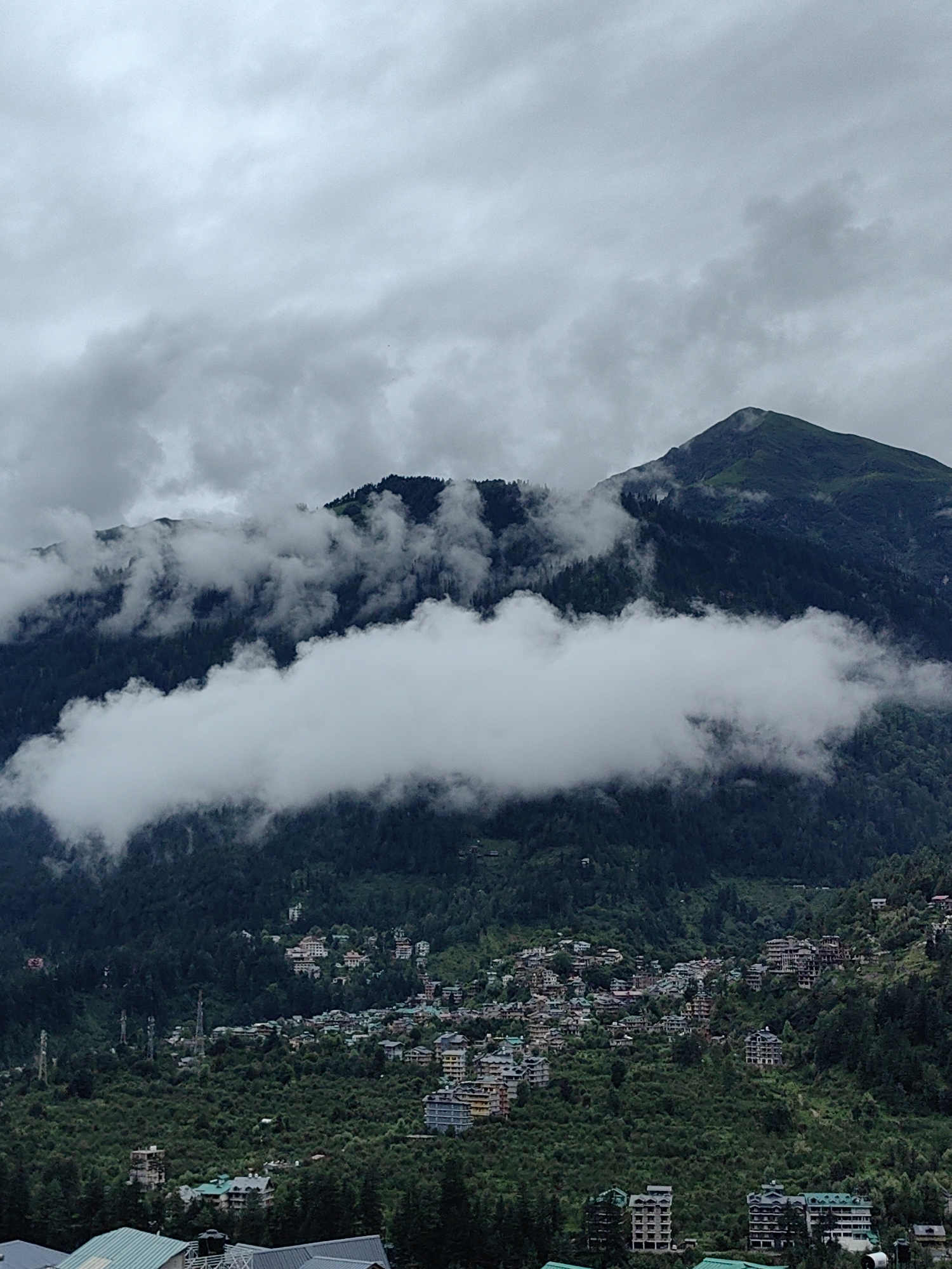 This is an aerial, full-frame shot of a mountain village in Vashisht, India. The sky is overcast with thick, gray clouds. A large, dark green mountain dominates the background, with patches of mist clinging to its upper slopes. A thick band of white clouds stretches horizontally across the middle of the image, partially obscuring the mountain. Below the clouds, the steep mountain slope is covered in dense, dark green pine trees. A village is nestled into the side of the mountain, with numerous buildings of varying sizes and colors, primarily white and pastel hues with some darker tiled roofs. The buildings are clustered together, following the contours of the land. Some are multi-story hotels or apartment complexes, while others appear to be smaller homes. Several tall, thin cell towers are visible among the trees in the mid-ground. The foreground shows the rooftops of several buildings with green metal roofs, suggesting the viewpoint is from a higher elevation looking down. The overall scene depicts a densely populated village set against a dramatic, cloudy mountain landscape.