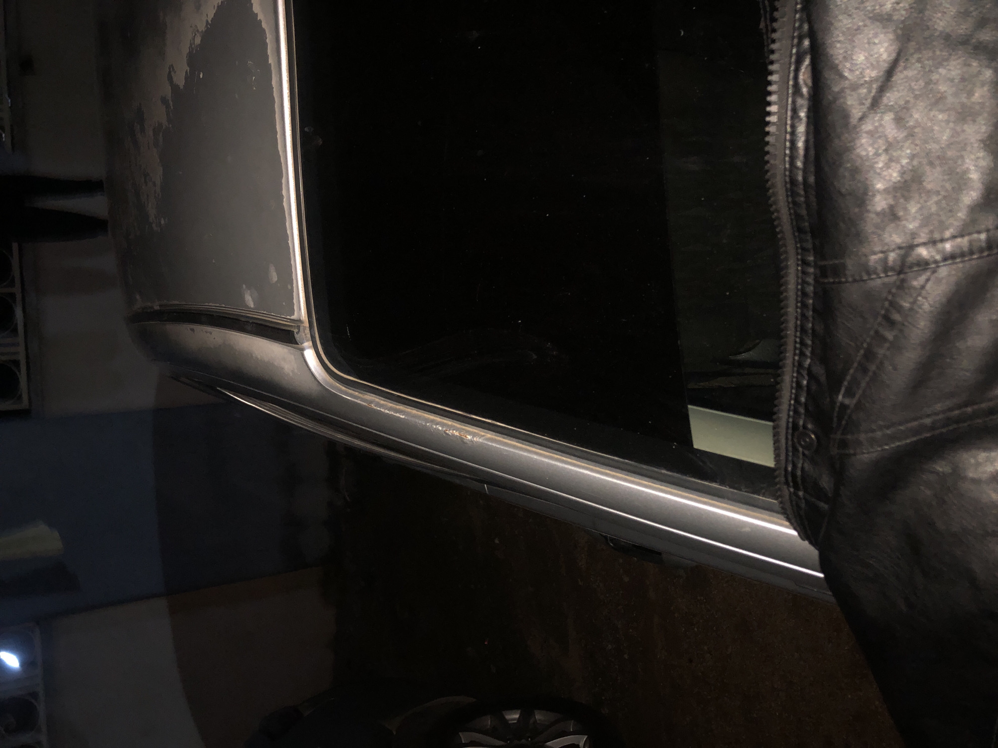 The image presents a nighttime outdoor scene in Unknown, Unknown. In the immediate foreground, the upper portion of a black leather jacket, displaying texture and a zipper, occupies the lower right. Directly behind the jacket, the roof and upper side frame of a light-colored sedan are visible. The car's roof exhibits extensive paint degradation and discoloration. Above the vehicle, from the knees down, a person wearing dark trousers is seen standing on a light-colored ledge or wall. To the far left of the car, a portion of a vehicle wheel with silver and black spokes is visible. The background consists of dark building facades or walls, with a distinct bright light source present in the upper left corner. The ground is dark and appears uneven.