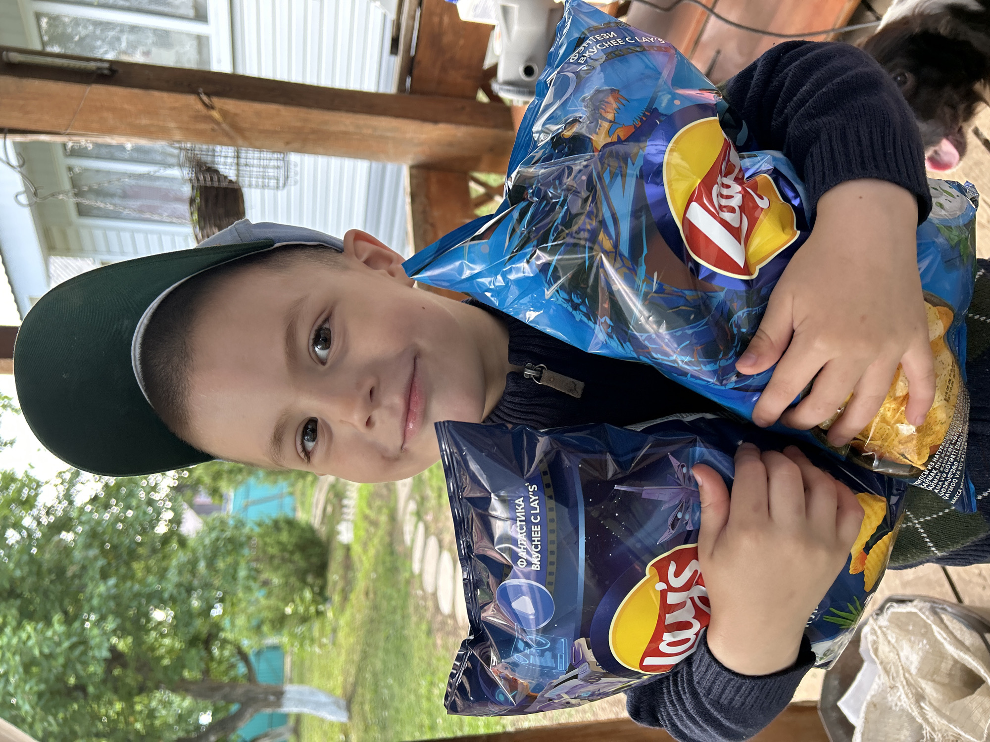 A young boy, approximately 4-6 years old, is depicted in a medium shot, smiling directly at the camera, in the city of Фоки, Russia. He wears a dark blue long-sleeved, ribbed knit sweater with a zippered collar and a dark green baseball cap with a light grey underside. His hands hold two large bags of Lay's potato chips.
The bag in his left hand is predominantly dark blue with a cosmic/futuristic theme, displaying a stylized spaceship and abstract patterns. Cyrillic text visible on this bag reads "ФАНТАСТИКА ВКУСНЕЕ С LAY'S," which translates to "FANTASTIC TASTES BETTER WITH LAY'S." The bag in his right hand is predominantly light blue with a fantasy theme, featuring a large, ornate dragon-like creature. Cyrillic text on this bag reads "ФЭНТЕЗИ ВКУСНЕЕ С LAY'S," translating to "FANTASY TASTES BETTER WITH LAY'S." Both bags prominently feature the red and yellow Lay's logo.
The setting appears to be an outdoor porch or veranda. A rustic wooden post of a structure is visible on the right, and behind it, a white building with horizontal siding and windows. A hanging basket is suspended from the structure. The background on the left features blurred green foliage from trees, one of which has a white-painted base. A bright turquoise fence is partially visible behind the trees, and stone stepping-stones are on the ground. In the bottom right corner, a portion of a brown and white dog's head with its mouth open and tongue visible is partially captured.