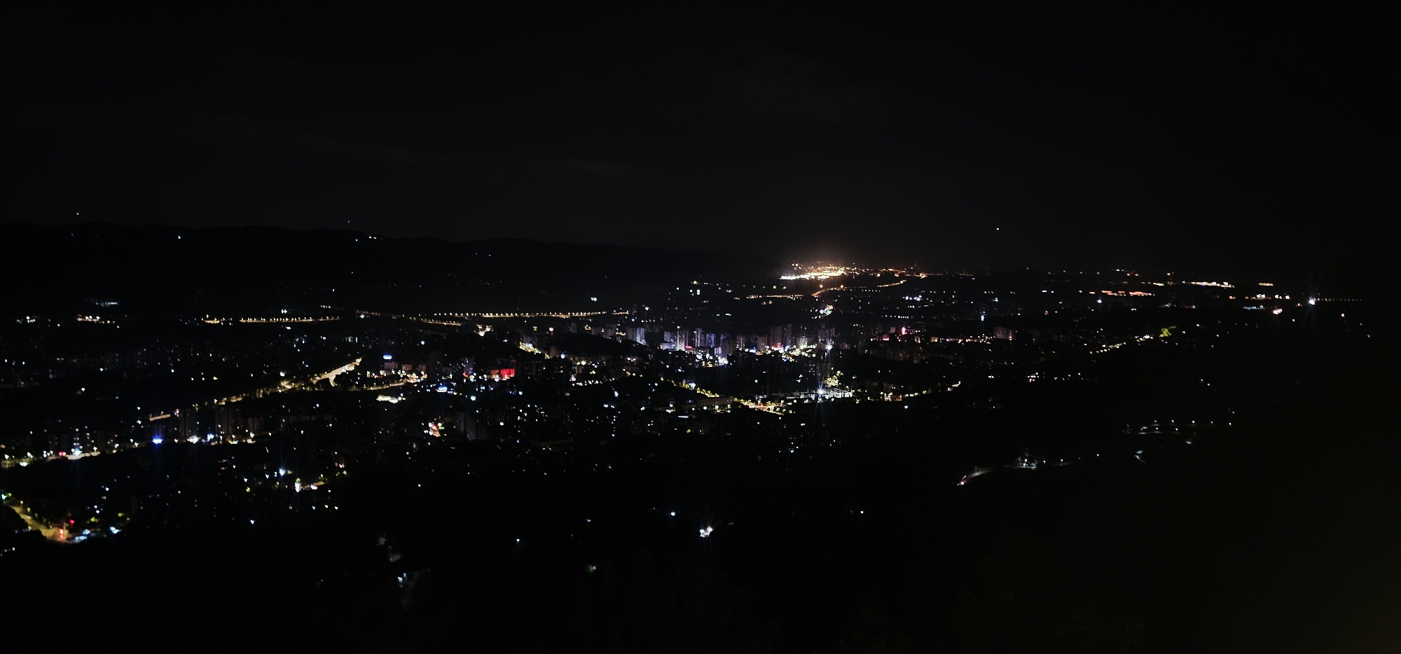 This is a panoramic nighttime photograph of the city of Chengjiang, China, taken from a high vantage point. The foreground is dark, with silhouetted hills and foliage, suggesting the viewer is on a mountainside overlooking the city. The city itself is illuminated by a vast array of lights, forming a tapestry of various colors and intensities. Tall buildings, likely apartment complexes or office towers, are visible as clusters of bright, rectangular lights, some appearing to be lit with white, red, and blue illumination. Roads are traced by lines of streetlights, and more scattered lights suggest residential areas and businesses. In the distance, a brighter, more concentrated area of illumination hints at a central business district or a major commercial hub within Chengjiang. The sky is dark, with a few faint points of light possibly representing stars or distant aircraft. The overall impression is one of a sprawling urban landscape actively alive with light under the cover of night.