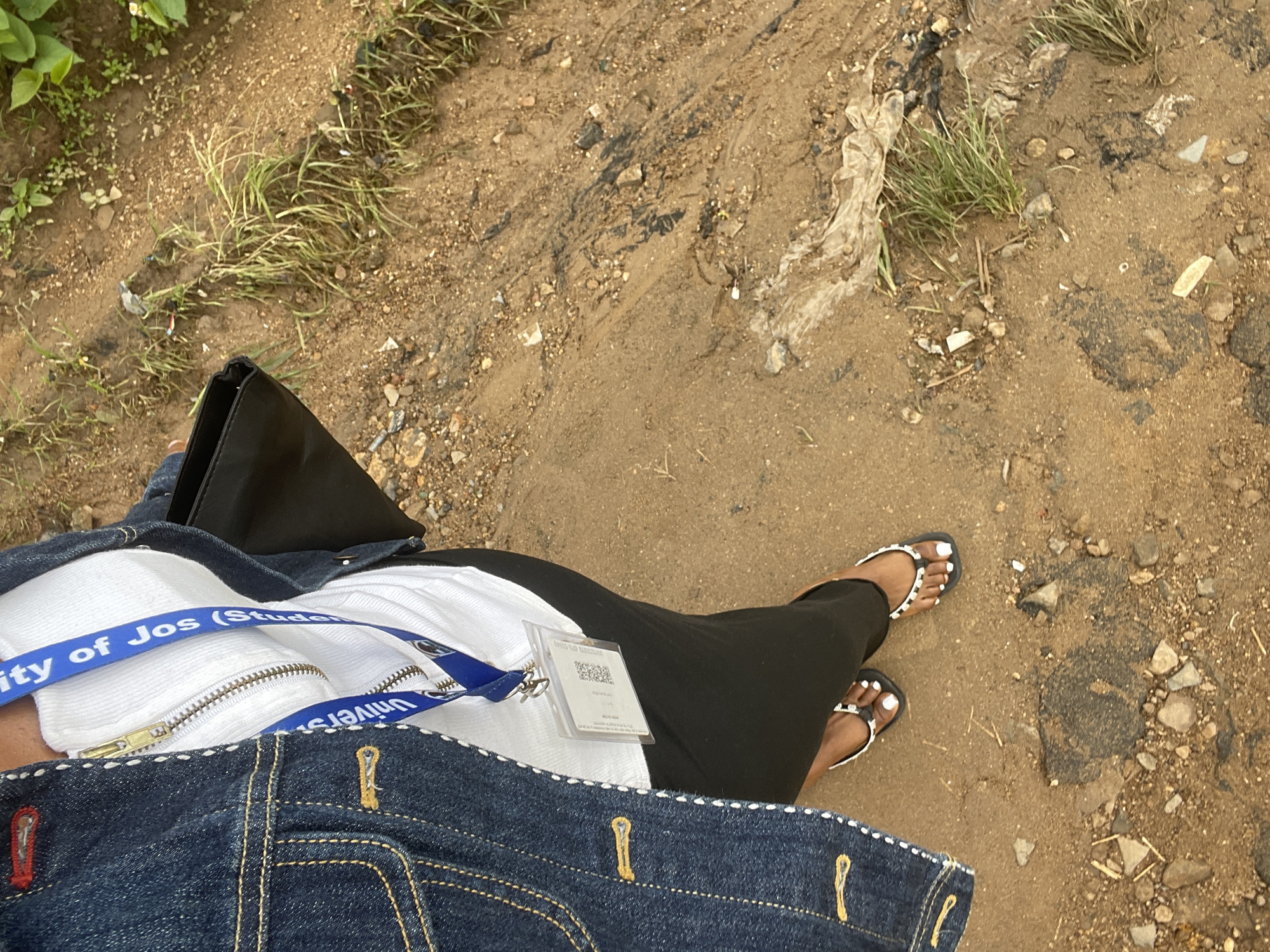 The image shows a close-up of a person's lower body, from the waist down, standing on a dirt path. The person is wearing a blue denim jacket, a white zippered top, and a black skirt. A blue lanyard with an ID card is visible around the person's neck. The person is wearing flip-flops. The ground is a mix of dirt, small rocks, and some sparse vegetation. The scene appears to be outdoors, possibly in a rural or semi-urban area. The environment is natural and appears somewhat unkempt, with some debris visible on the ground. The lighting suggests daylight. The context of Rafin Par, Nigeria is not explicitly depicted in the image itself, but is provided as an external detail.
