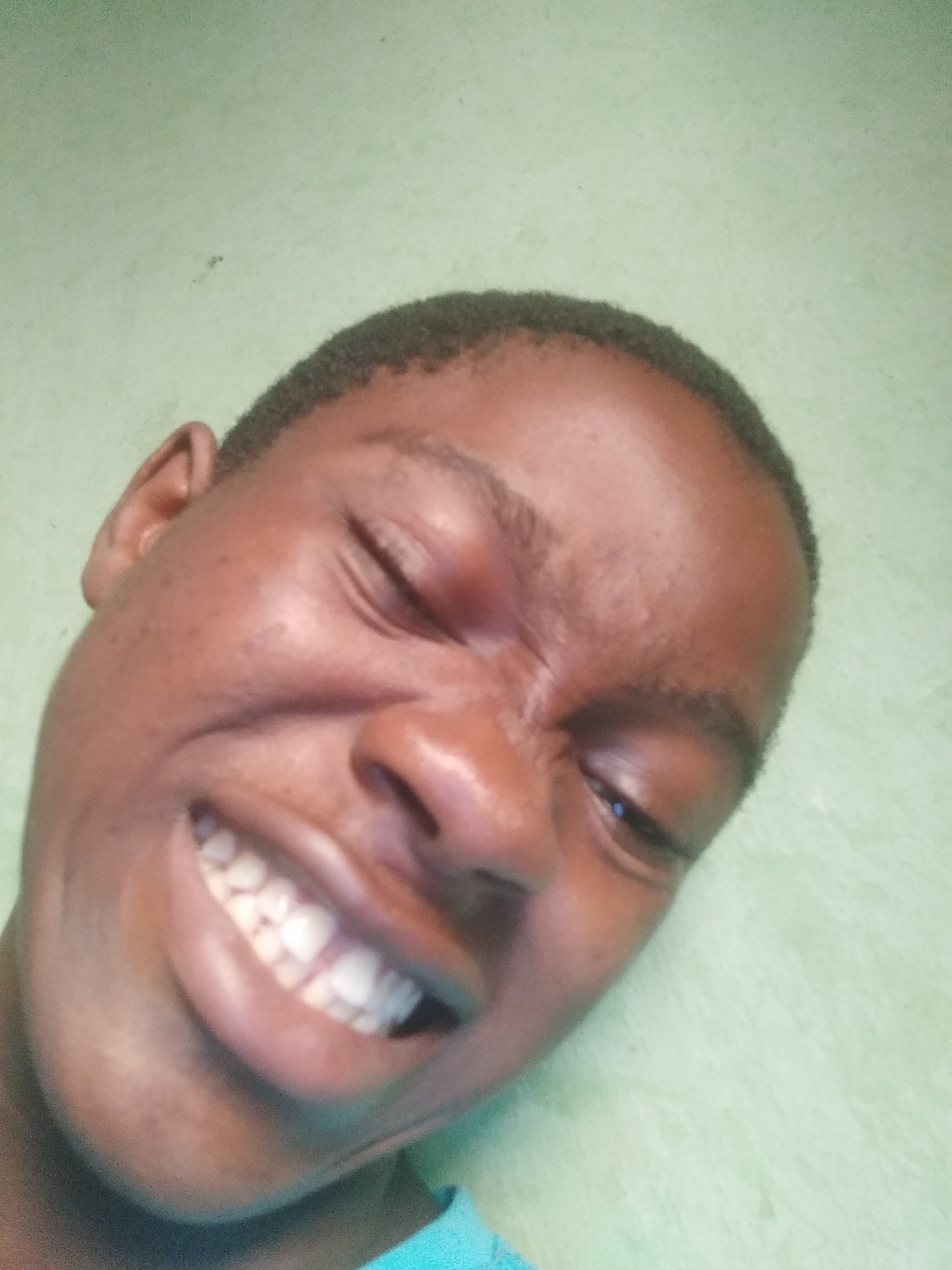 A close-up selfie captures a young Black man with a shaved haircut. His eyes are partially closed, and he has a wide, toothy grin. The background is a pale green wall. The specific location within Shangisha, Nigeria, cannot be determined from the image.