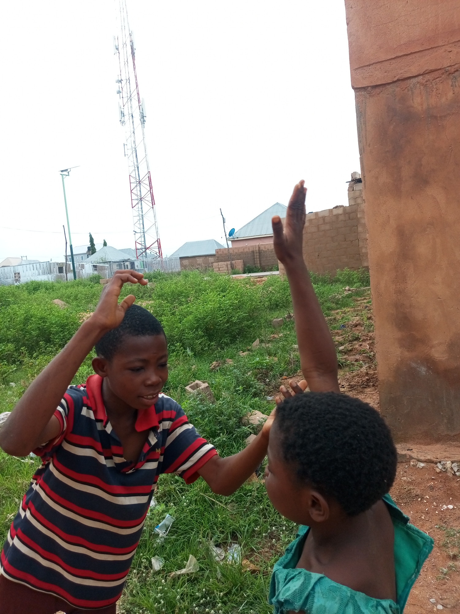The image depicts two young children engaged in an activity outdoors in Tempure, Nigeria. A boy, facing left and wearing a striped polo shirt, is positioned in the foreground. His right hand is raised, with fingers curled as if in mid-action. To his right, a younger child, facing away from the camera, is also raising their right arm, their hand out of frame. The environment suggests a somewhat undeveloped urban or semi-urban area. In the background, a tall telecommunications tower dominates the skyline. Various low-rise buildings, some under construction with exposed brickwork, are visible, along with fences and scattered debris. The ground is covered with green vegetation and patches of dirt. The sky is overcast, providing diffused light.