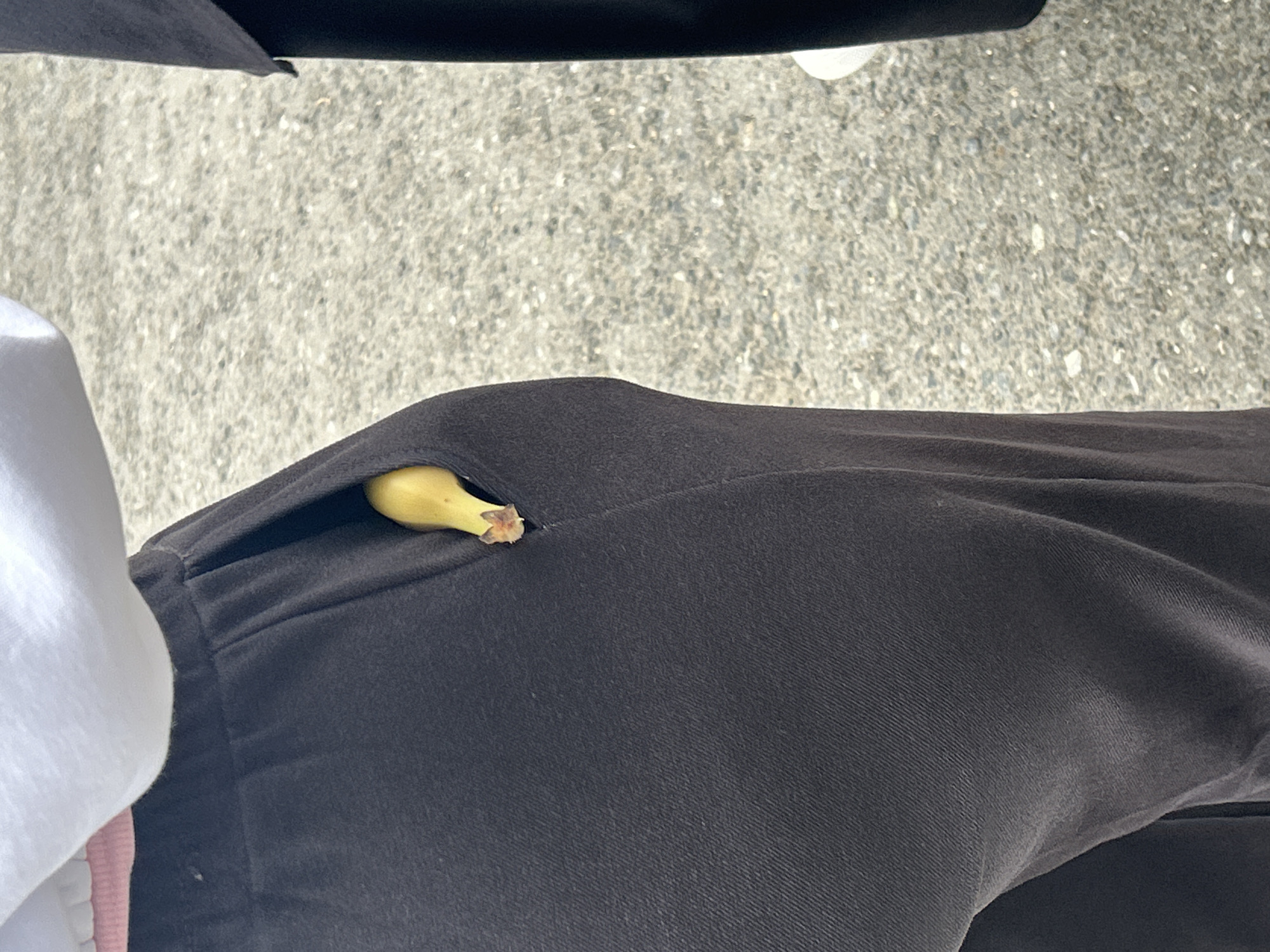 The image shows a close-up view of a person's dark fabric pants with a small piece of a yellow banana protruding from the pocket. The background appears to be asphalt, suggesting an outdoor urban environment. The image does not contain any information that can be used to determine the location in the city of Perm, Russia.