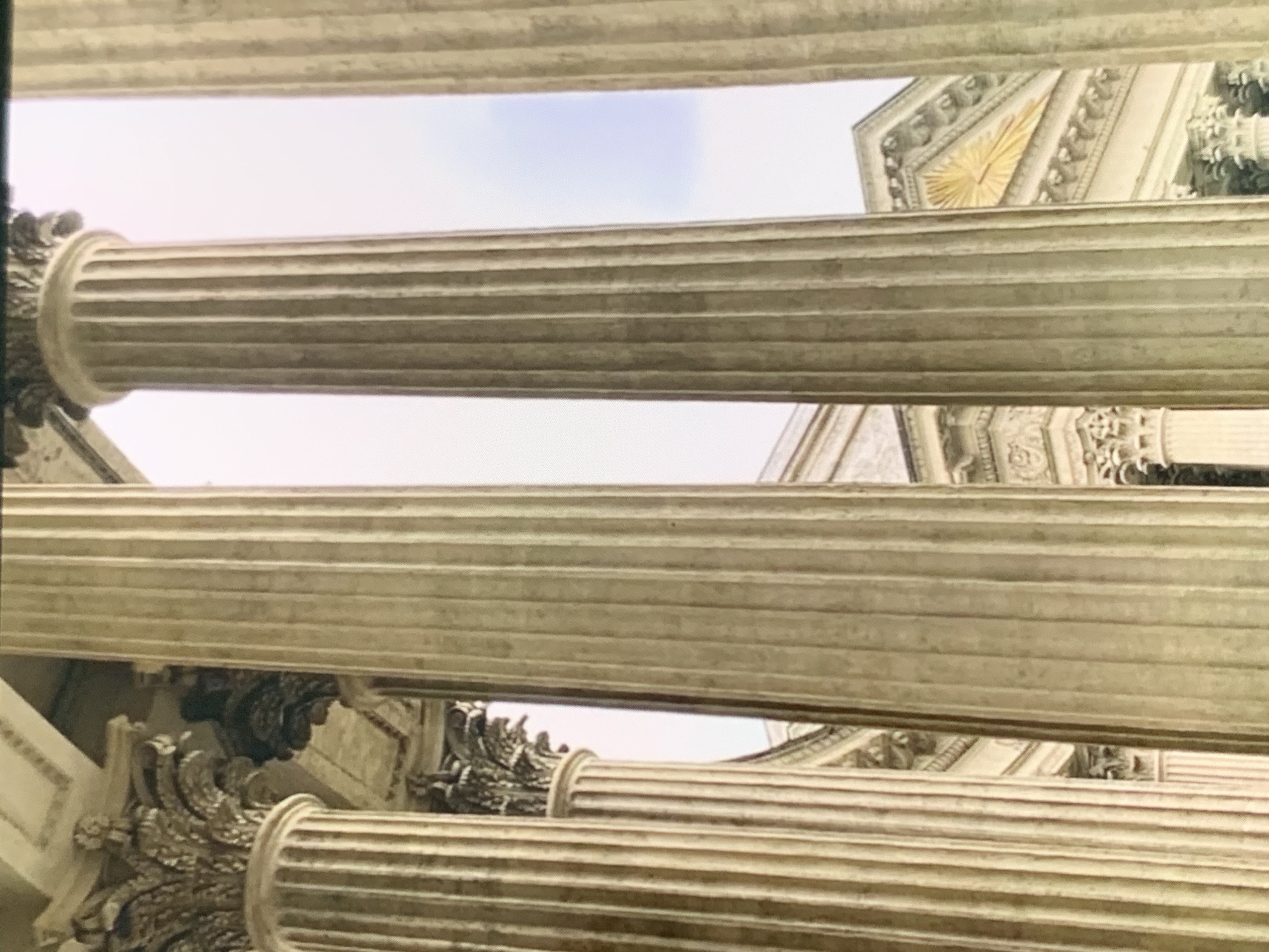 The image depicts a view through a colonnade of classical-style columns, with architectural elements of a grand building visible in the background. The columns are fluted and made of a light-colored stone, possibly marble or limestone. They are positioned in the foreground and mid-ground, creating a sense of depth and framing the scene.
In the background, behind the columns, are parts of a building with ornate detailing. There are visible pediments, cornices, and decorative carvings. One prominent feature is a triangular pediment adorned with what appears to be a golden sunburst or radiant motif. The architecture suggests a historical or public building, possibly a museum, government institution, or a grand estate.
The sky is visible in the upper portion of the image, appearing overcast or with diffused light, as the colors are muted. The overall lighting is soft, indicating it could be daytime, but not direct sunlight. There are no people or animals visible in the image. The perspective is from a low angle, looking upwards slightly towards the architecture.
The building depicted is the Stavropol State Museum of Local Lore named after G.N. Prozritelev and G.K. Prave. It is a prominent architectural landmark in Stavropol, Russia. The museum is known for its neoclassical style, characterized by its grand columns and detailed ornamentation, as seen in the image. The specific viewpoint seems to be from the area of the museum's entrance or a nearby courtyard, looking towards the facade.