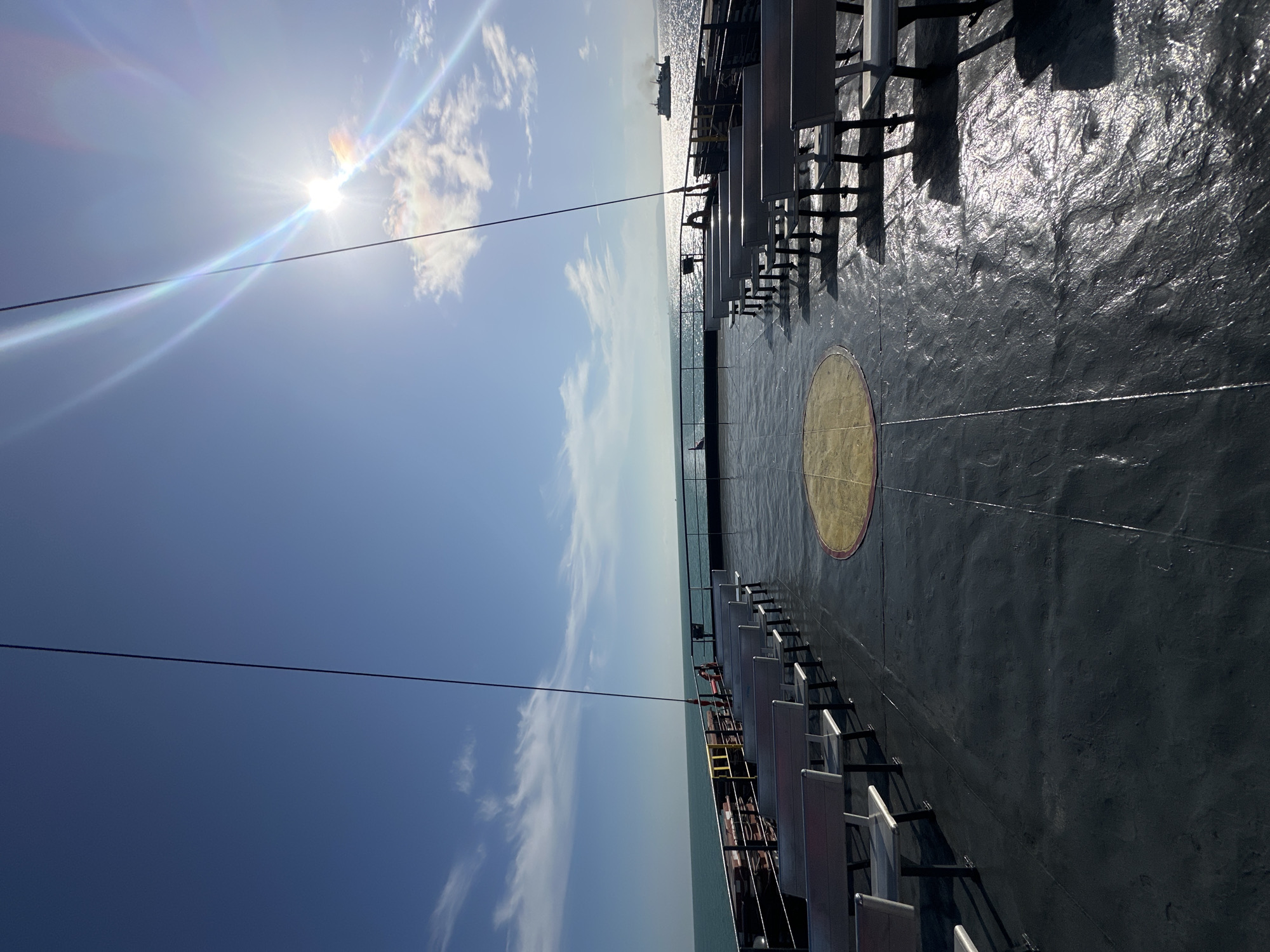The image shows the deck of a ferry in the city of Don Sak, Thailand, on a clear, sunny day. Rows of empty, silver benches are arranged on the dark gray deck, facing the ocean. A circular yellow and red marking is visible on the deck in the center. The turquoise ocean stretches to the horizon under a bright blue sky. The sun is high in the sky, causing lens flare and casting long shadows from the benches. In the distance, a cargo ship can be seen on the water. The overall scene conveys a sense of calm and anticipation, as passengers await their journey.