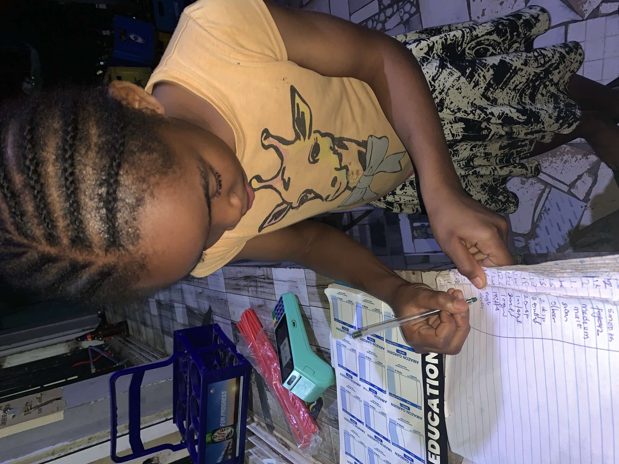 A person is writing on a notebook with a pen. The notebook is resting on a stack of forms with "Amazon Garden Education" printed on them. Beside the notebook are a handheld POS device, a package of red straws, and a blue plastic crate labeled "Life FOR PROGRESS". The person appears to be inside a building. The OCR text includes "Garden Amazon Garden," "Total N," and "Die FOR PROGRESS". The environment, specifically "New Stadium, Nigeria," is implied. The notebook contains handwritten text, including "smooth", "medium", "malt", "Swan", "Olver", "2 small", "Eagle", "Double".