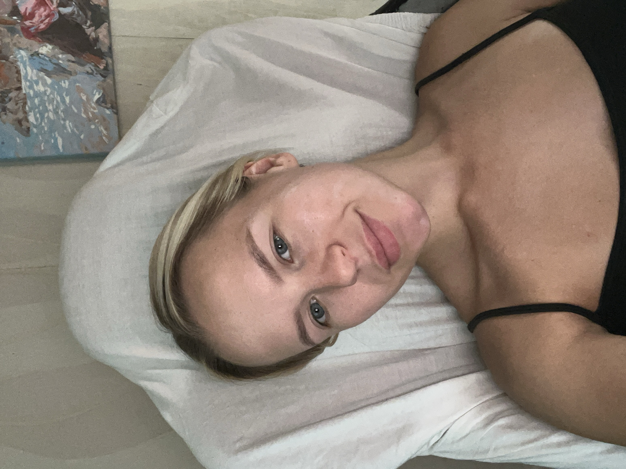 A young Caucasian woman with blonde hair and blue eyes is lying down on a white sheet, looking directly at the camera. She is wearing a black spaghetti-strap top. Her face is partially illuminated by natural light, highlighting her fair skin and rosy cheeks. A painting with a coastal landscape is visible on the wall in the background. The setting appears to be a room in an unknown location within a city.