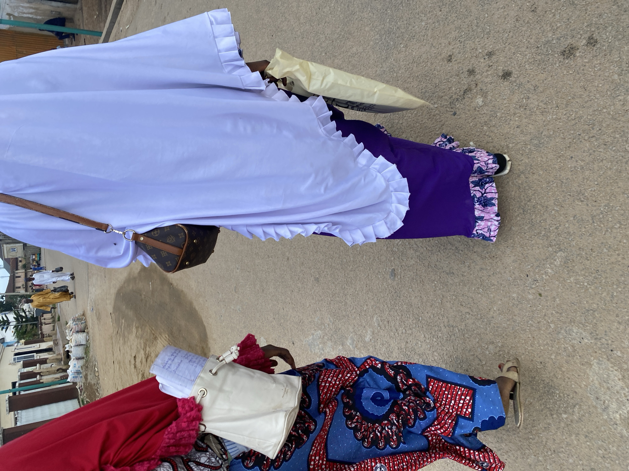 Two individuals, likely women, are walking on a paved street in Jos, Nigeria. The person on the left is wearing a red headscarf and a vibrant blue and red patterned skirt. They are carrying a white bag with a pink fringe. The person on the right is wearing a white head covering and a long purple dress with a ruffled hem. They are carrying a light-colored shopping bag. In the background, a man in a yellow garment is walking away from the camera, followed by another person in a white outfit. Bags of cement are visible stacked on the side of the road, and there are buildings and some vegetation in the distance, indicative of a semi-urban or residential area. The lighting suggests it is daytime, likely mid-morning or afternoon.