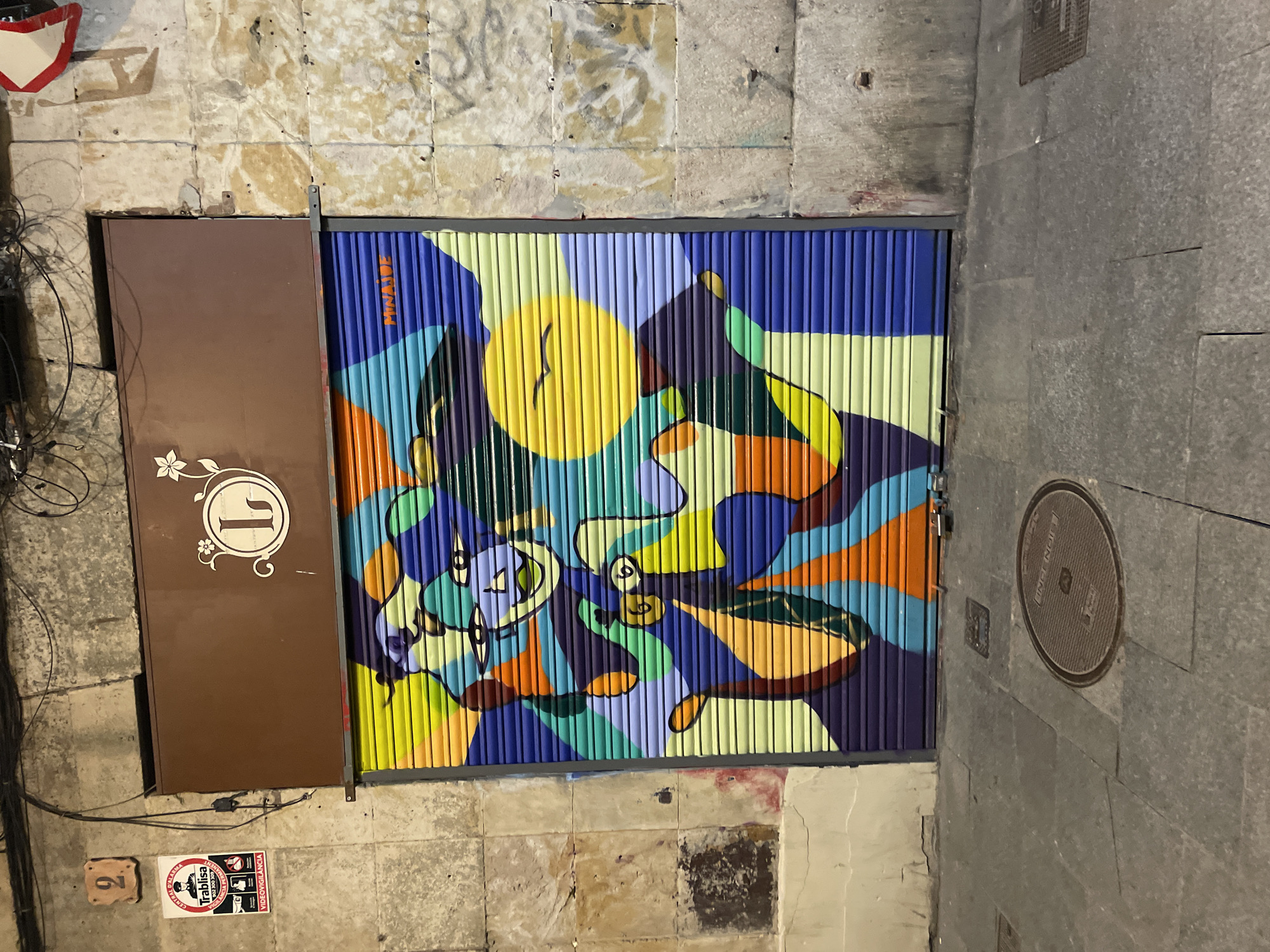 The image captures a nighttime street scene in Barcelona, Spain, centered on a storefront with a distinctive security shutter. Above the shutter, a brown rectangular sign displays a white circular logo featuring the letter "L" adorned with a floral vine motif. The metal security shutter itself is painted with a vibrant, abstract mural. The mural incorporates multiple colors including shades of blue, green, yellow, orange, and purple, depicting stylized faces, a prominent yellow sun-like disc with two internal strokes resembling a bird, and abstract shapes. The artist's signature, "MINAJUE," is visible in orange text on a blue section of the shutter.
To the left of the storefront, a beige stone wall features a ceramic tile with the number "2" and a rectangular sticker. The sticker, bearing the "Trablisha" logo, reads "CENTRAL D'ALARMA" and "VIDEOCVIGILANCIA," confirming the Catalan linguistic context of Barcelona. Above, numerous tangled electrical wires and cables run along the wall. The rough-textured stone walls flanking the storefront exhibit wear, stains, and minor graffiti. A partially visible red and white triangular sign is attached to the upper right wall. The foreground consists of a grey paved sidewalk or street, featuring a circular metal manhole cover with "INGENIERIA" text and a rectangular grate. The scene is illuminated by artificial light, casting soft shadows.