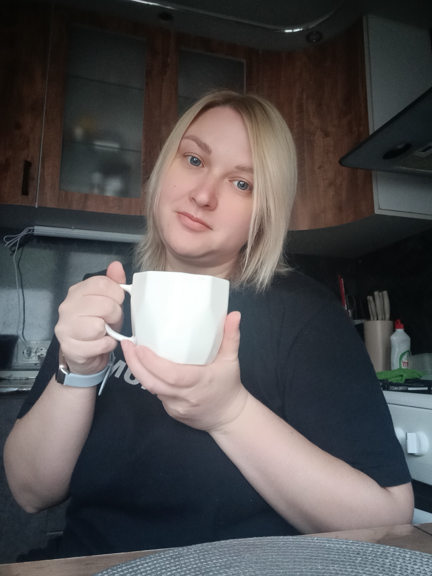A woman with light blonde hair and blue eyes holds a white, angular coffee mug in her kitchen. She is wearing a black t-shirt and a smartwatch on her left wrist. The kitchen features wooden cabinets, a stove, and various kitchen utensils. The location is Chaikovsky, Russia.