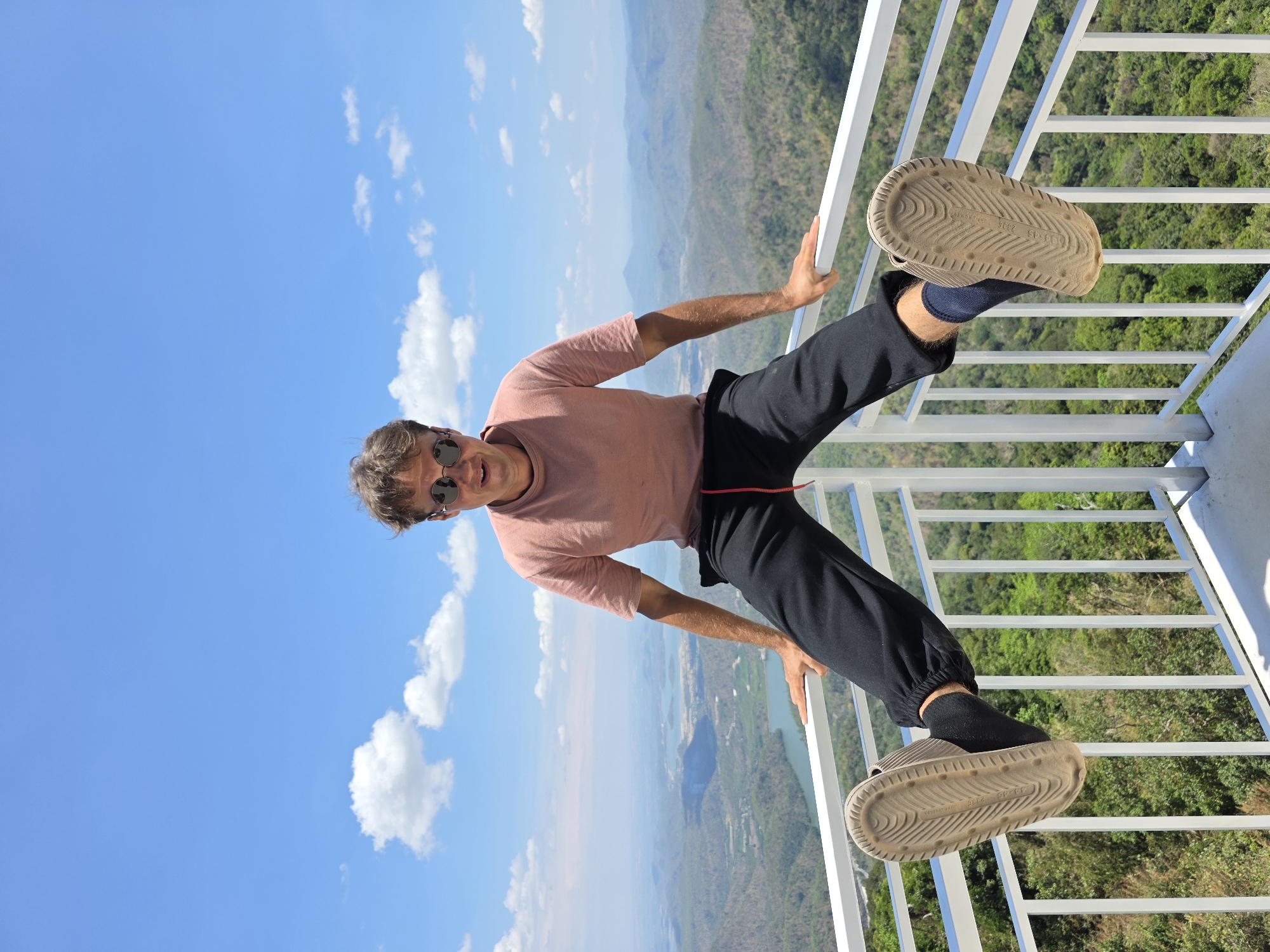 The image displays a man posing playfully on an elevated outdoor observation deck, offering a panoramic view of a vast landscape. The man, with short, curly brown hair and sunglasses, is wearing a light reddish-brown t-shirt, black joggers, black socks, and light brown slides. He is smiling slightly at the camera, holding onto a white metal railing with both hands, and has one leg bent with his right foot resting on the railing.
Behind him, the scene unfolds into an expansive vista of green hills and mountains, suggesting a forested or natural terrain. In the distance, a body of water, possibly a river or lake, is visible winding through the landscape. The sky above is a clear, vibrant blue with scattered white cumulus clouds, indicating bright daylight, likely midday or early afternoon. The bright lighting illuminates the scene, highlighting the textures of the landscape and the man's attire.
On the sole of the man's right slide, visible text includes "MADE IN CHINA" and "235", which likely denotes a shoe size. Other faint markings and a textured pattern are also present on the sole. The setting and the man's pose suggest he is enjoying a scenic viewpoint, consistent with an overlook in a picturesque region like Nam Hải, Vietnam.