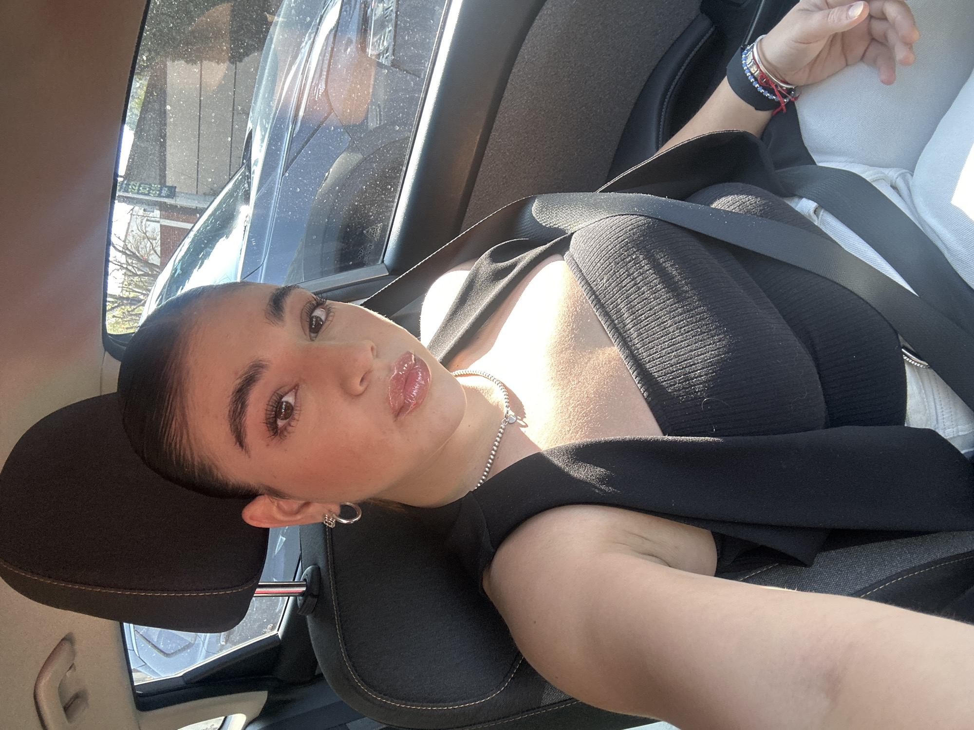 A young woman is photographed from a low angle inside a car. She is fair-skinned, wearing a black top, a silver necklace, and a black blazer or coat draped over her shoulders. She is wearing a seatbelt. Her hair is slicked back. She has bracelets on her left wrist and a silver earring in her left ear. The interior of the car is visible, including the headrest, the window, and the seat. Through the car window, a street with buildings is partially visible, which may be located in the city of Puebla, Mexico.