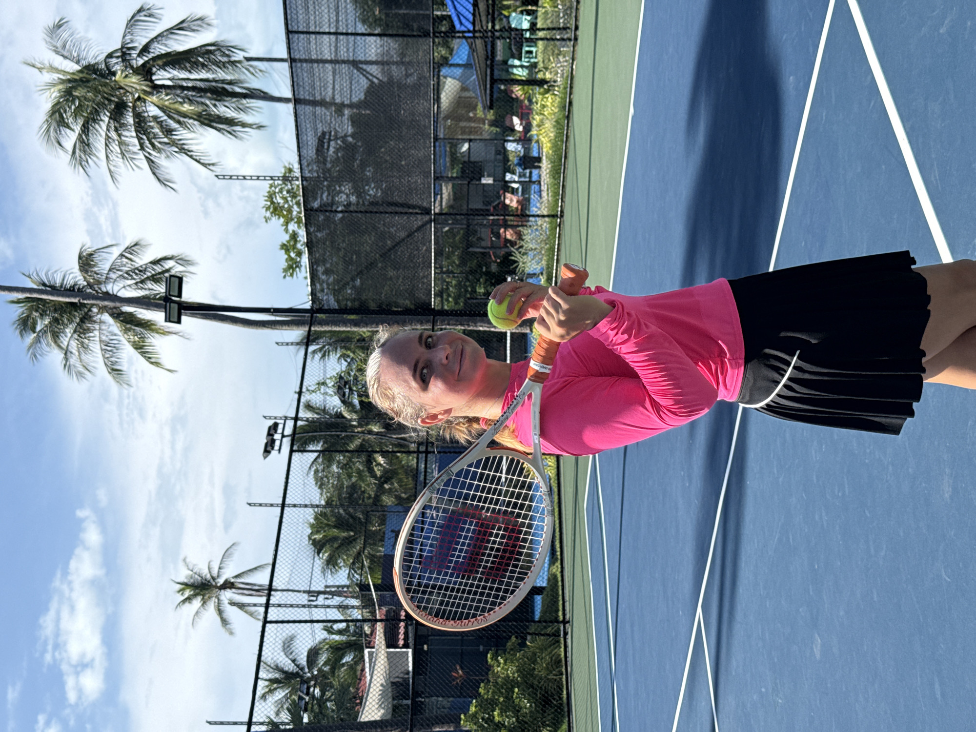 In Baan Plai Laem, Thailand, on a bright, sunny day with a partially cloudy sky, a young woman with blonde hair pulled back stands on a blue tennis court. She is wearing a vibrant pink long-sleeved shirt and a black tennis skirt. She is holding a tennis racket horizontally over her shoulder with her right hand and a tennis ball in her left hand. The tennis racket has a white frame and strings, with orange accents on the handle. The tennis court has white lines marking its boundaries. In the background, a black chain-link fence encloses the court, beyond which are palm trees, lush greenery, and some buildings. The scene suggests a recreational setting, possibly a resort or a sports complex.