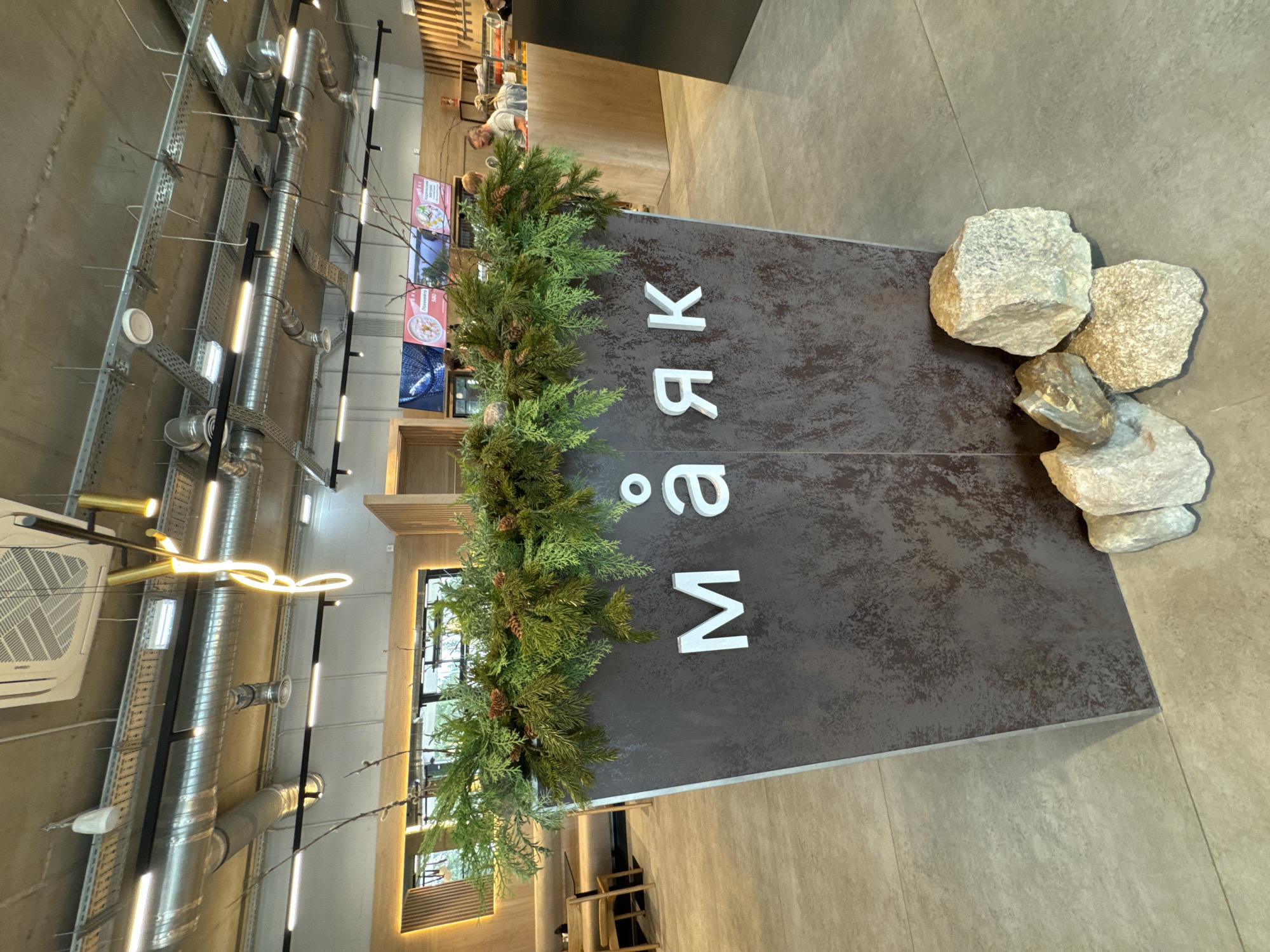 This image shows the interior of a modern cafe or restaurant. In the foreground, a large, dark brown planter box with a textured, metallic finish is decorated with evergreen branches and small pinecones. The word "МАЯК" (Mayak), meaning "lighthouse" in Russian, is prominently displayed on the front of the planter in large, white, three-dimensional letters. The planter is surrounded by several natural rocks of varying sizes, placed on a neutral-colored tiled floor.
The background reveals more of the establishment's interior. To the left, there are dining tables and chairs, suggesting a seating area. Above the planter, a wooden paneling wall is visible, and further back, screens display menu items or advertisements. The ceiling features exposed ventilation systems, industrial lighting fixtures, and a modern, decorative neon light. In the mid-ground, several people are visible near a counter or service area, including a man interacting with what appears to be a cashier. The overall atmosphere is contemporary and clean. The image does not contain any information that places it in the city of Yakkima, Russia.