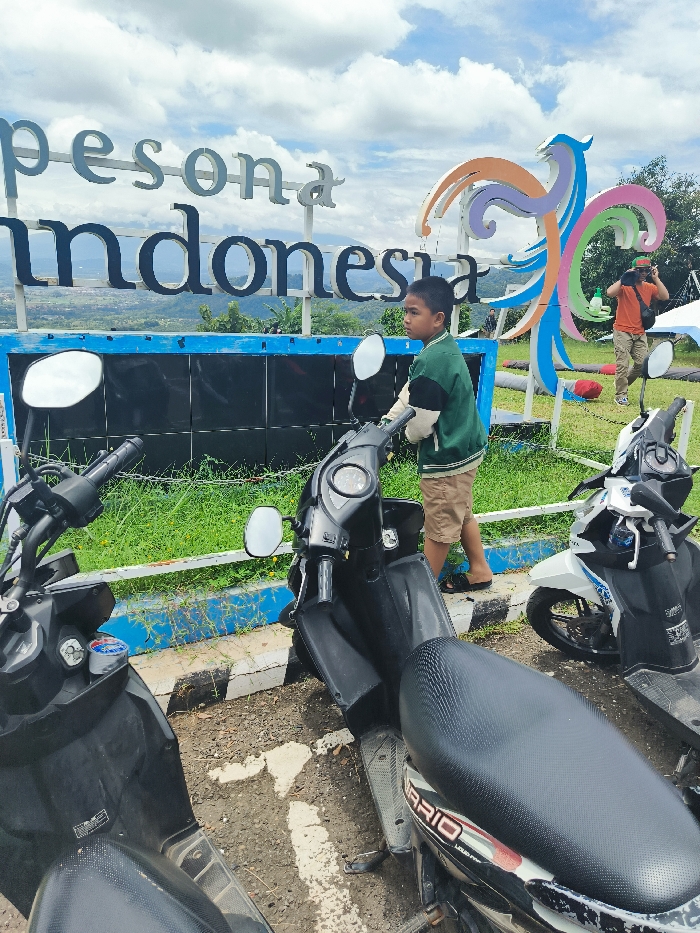 The scene is a parking lot in the city of Majalengka, Indonesia. The coordinates are -6.846 latitude and 108.204 longitude. There is a colorful sign that reads "pesona indonesia" with a large, abstract, colorful sculpture of a bird-like figure. A young boy in a green shirt and brown shorts is looking at a black scooter parked in front of the sign. He is standing on the edge of the parking lot, and the scooter is directly in front of him. Two other scooters, a black one and a white one, are in the background of the scene. A man in a blue shirt and black pants is sitting on the white scooter. The black scooter is in the foreground, and the white scooter is behind it. The man on the white scooter is looking toward the left of the image. Behind the scooters is a large, black, metal structure with a blue and white painted frame. The structure has a large, rectangular, black panel on it. There are a few green plants in front of the structure. The sky is blue and there are a few white clouds in the sky.