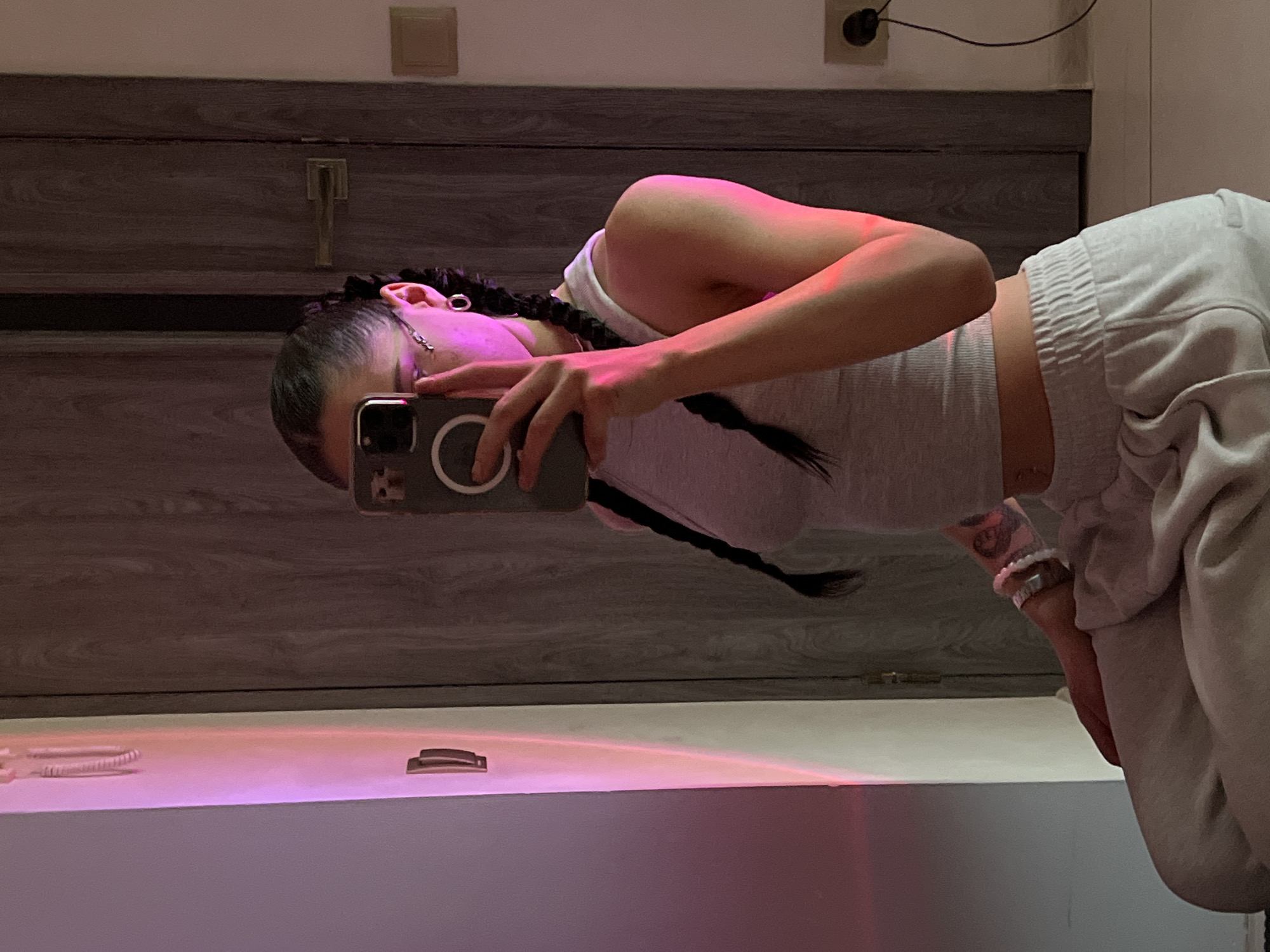 The image shows a young woman taking a selfie in a room. She is wearing a grey tank top and light grey sweatpants. Her hair is styled in two braids. She is holding a smartphone in her right hand, partially obscuring her face. A pink light is reflecting on her left arm and the wall behind her. The background includes a wooden door and an electrical outlet. There are no visible landmarks or specific details that would indicate the location is in Ternopil, Ukraine.