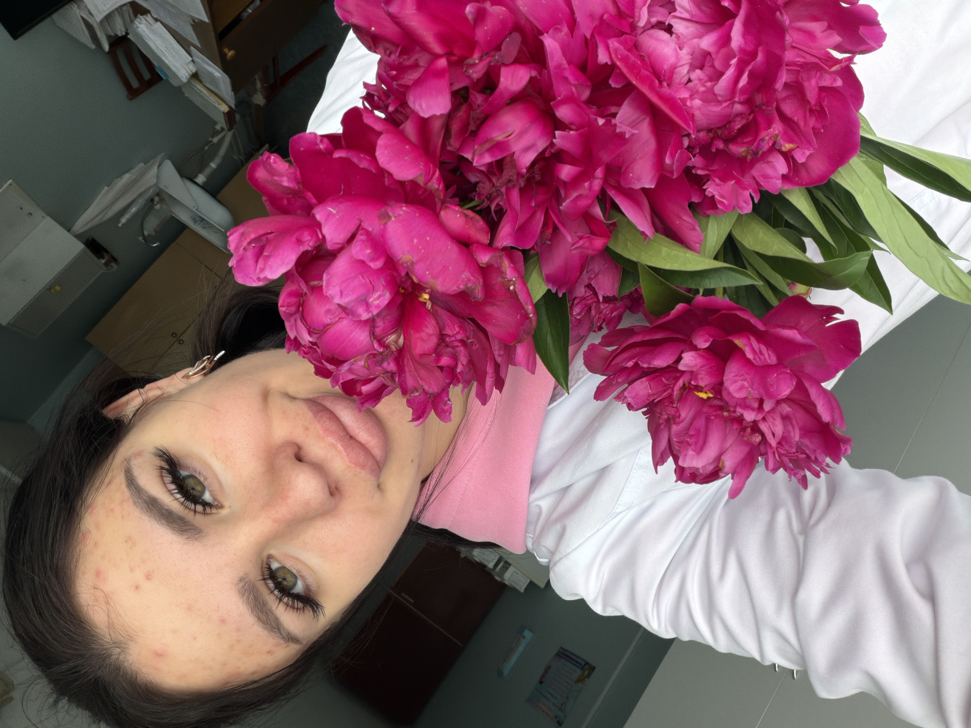 The image depicts a young woman in a white lab coat holding a bouquet of vibrant pink peonies. She is positioned in the foreground, with the flowers prominently displayed. Her face is turned towards the camera, and she appears to be taking a selfie. The background is blurred but suggests an indoor environment, possibly a medical facility, with cabinets and medical supplies visible. The woman has dark hair, is wearing dramatic false eyelashes, and has noticeable acne on her forehead and cheeks. The lighting is bright, casting a slight shadow on her face. The overall impression is one of a casual, personal moment captured within a professional setting. The image is a photograph, not a painting or drawing. The image does not contain any discernible information about the city of Chumna, Russia.