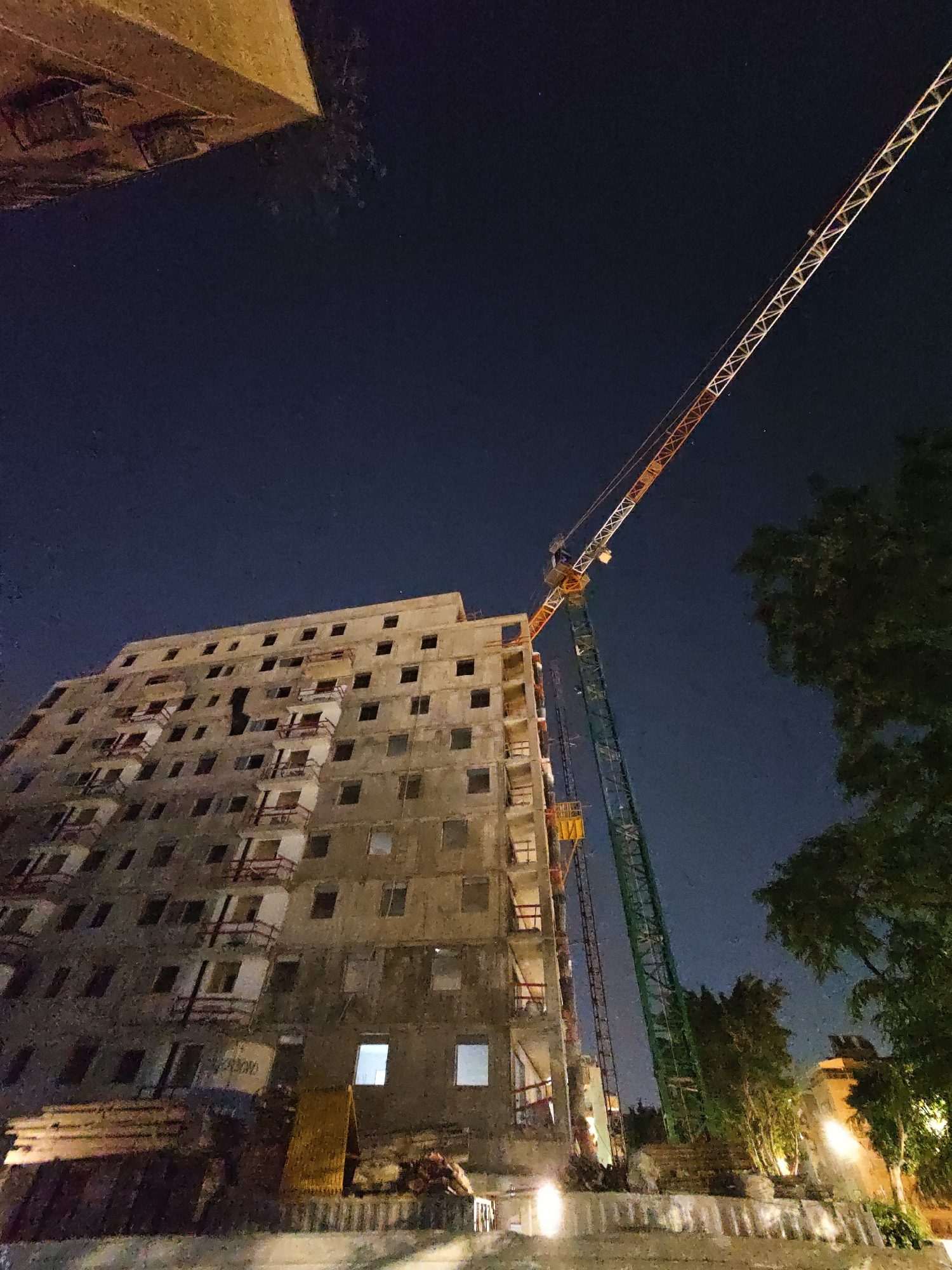 A low-angle, night-time photograph captures a construction site in Petah Tikva, Israel. Dominating the scene is a multi-story, unfinished apartment building with numerous open window frames and scaffolding. A tall construction crane, painted in sections of orange and green, extends diagonally across the dark night sky, its boom reaching high above the building. The sky is dark blue, suggesting nighttime. In the foreground, construction materials, possibly wood planks and debris, are piled near the base of the building. Trees with lush foliage are visible to the right of the building, partially obscuring the night sky. The scene is illuminated by artificial lights, casting a warm glow on some parts of the construction site and the building. No people are visible in the frame.