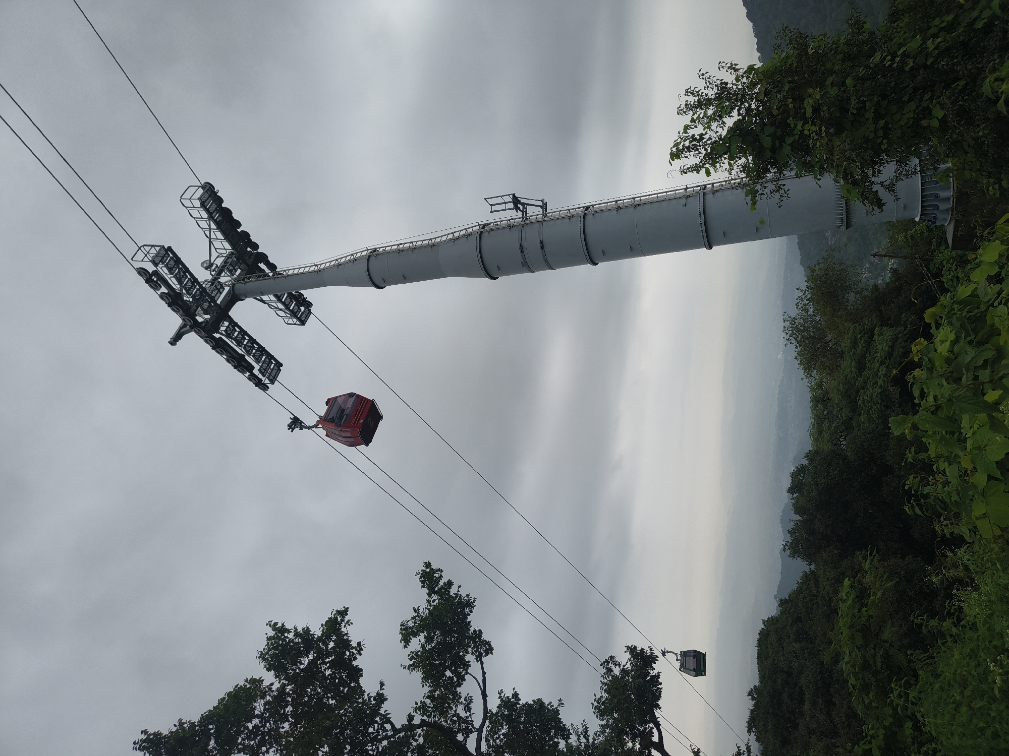 An image shows a ski lift with two gondolas, one red and one green, suspended on cables against a cloudy sky. The ski lift tower is a tall, gray structure with ladders and platforms. Trees and foliage are visible at the bottom of the image, with a hazy landscape in the background. The location is identified as Unknown, Unknown.