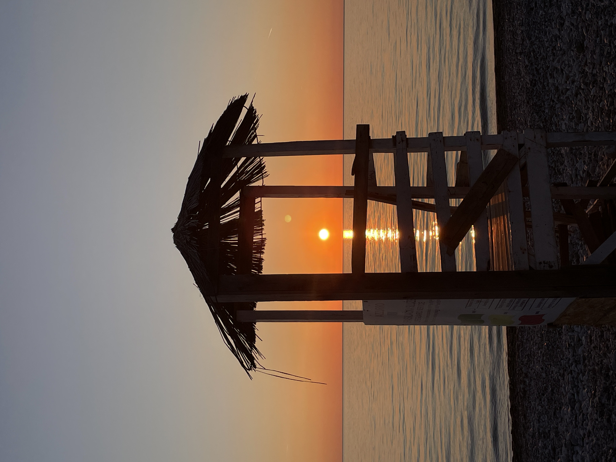 The image depicts a scene at sunset in Bar, Montenegro. The composition is dominated by a partially obscured lifeguard stand with a thatched roof in the foreground. The sun is positioned directly behind the stand, creating a silhouette effect and causing a bright reflection on the water's surface. The sky transitions from a light blue to an orange hue as the sun sets. The beach appears to be made of pebbles or small rocks. The water is calm, with subtle ripples reflecting the sunlight. A sign is attached to the side of the lifeguard stand, but the text is illegible.