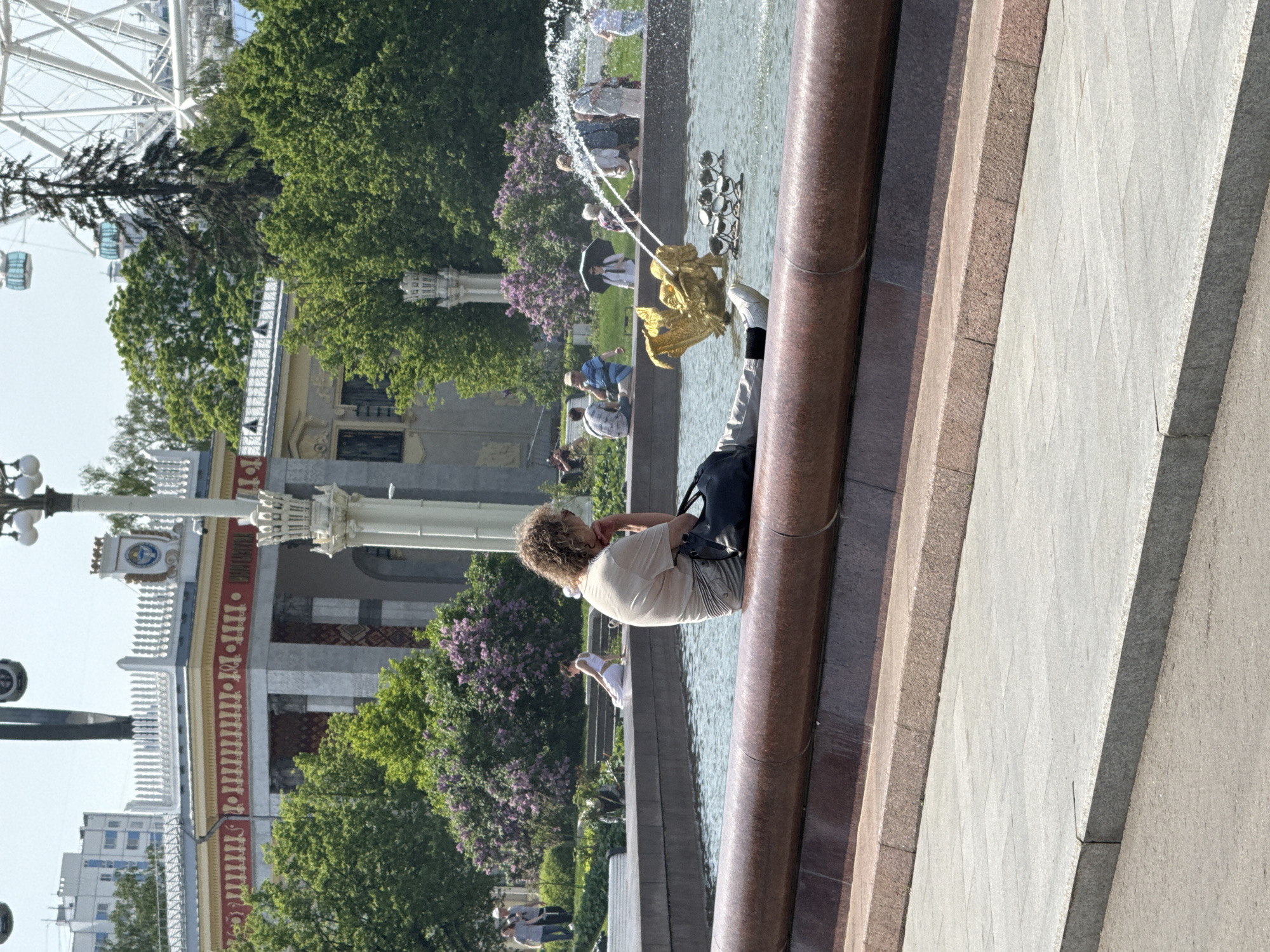 The image depicts a fountain in what appears to be a park setting in Moscow, Russia. A woman with curly hair is sitting on the edge of the fountain, with a black bag on her shoulder. People are visible in the background, some standing near the fountain and others walking in the park. Trees and flowering bushes are also visible. A Ferris wheel can be seen in the distance. A building with a white facade and red detailing is to the left, indicative of classical Russian architecture. The picture is taken at an angle, showing the stone work of the fountain and sidewalk.