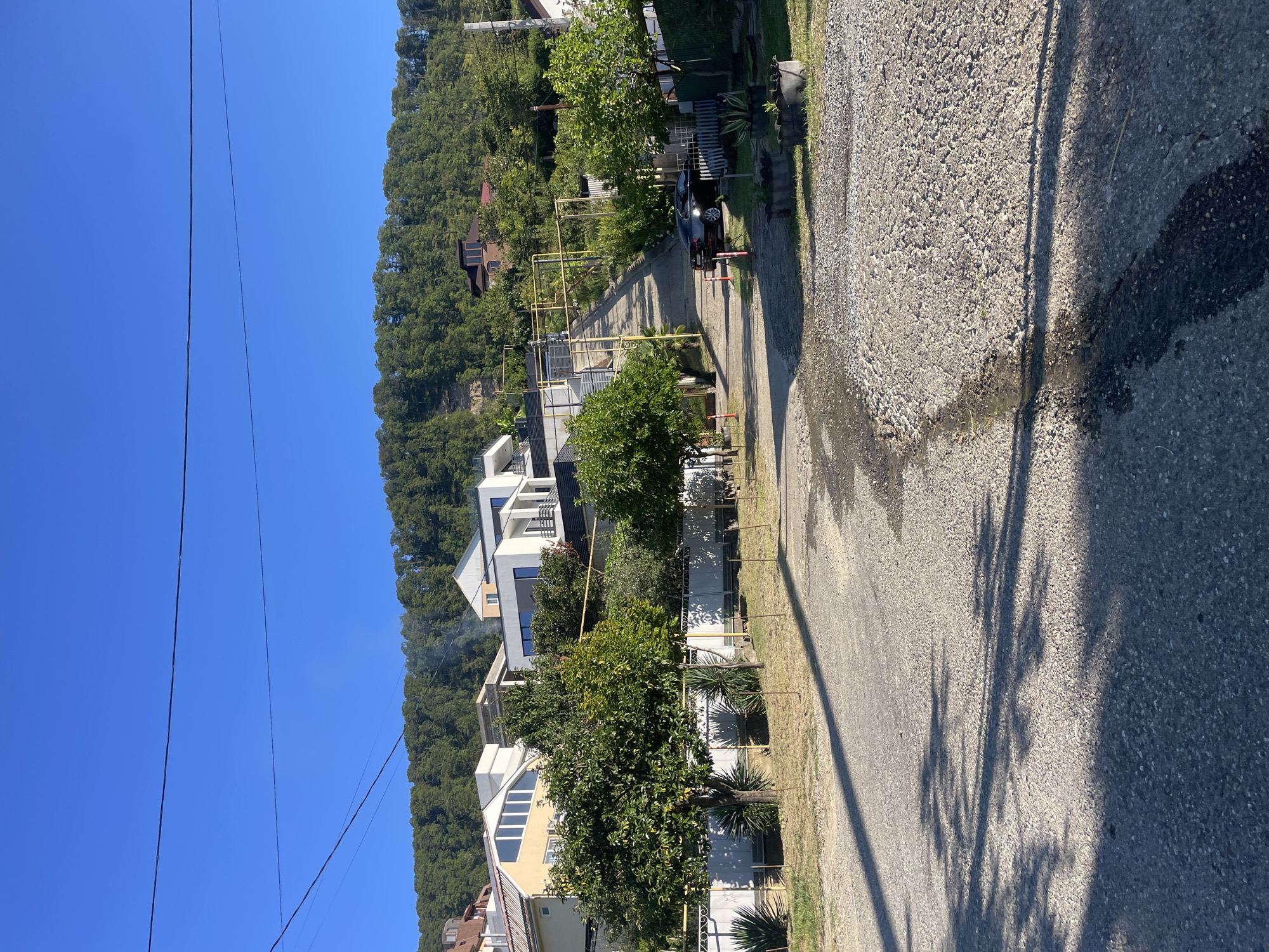 The image captures a street scene in Baranovka, Russia, under a clear blue sky. The road, paved with asphalt and gravel, winds uphill. On the left side of the road, modern white and yellow houses with large windows are visible, set against a backdrop of lush green trees. A white fence runs along the property line. On the right side, a steep asphalt incline leads further up the hill, flanked by greenery and more houses. A black car is parked on the right side of the uphill road. The overall environment suggests a residential area with a hilly terrain, typical of a coastal or mountainous region.