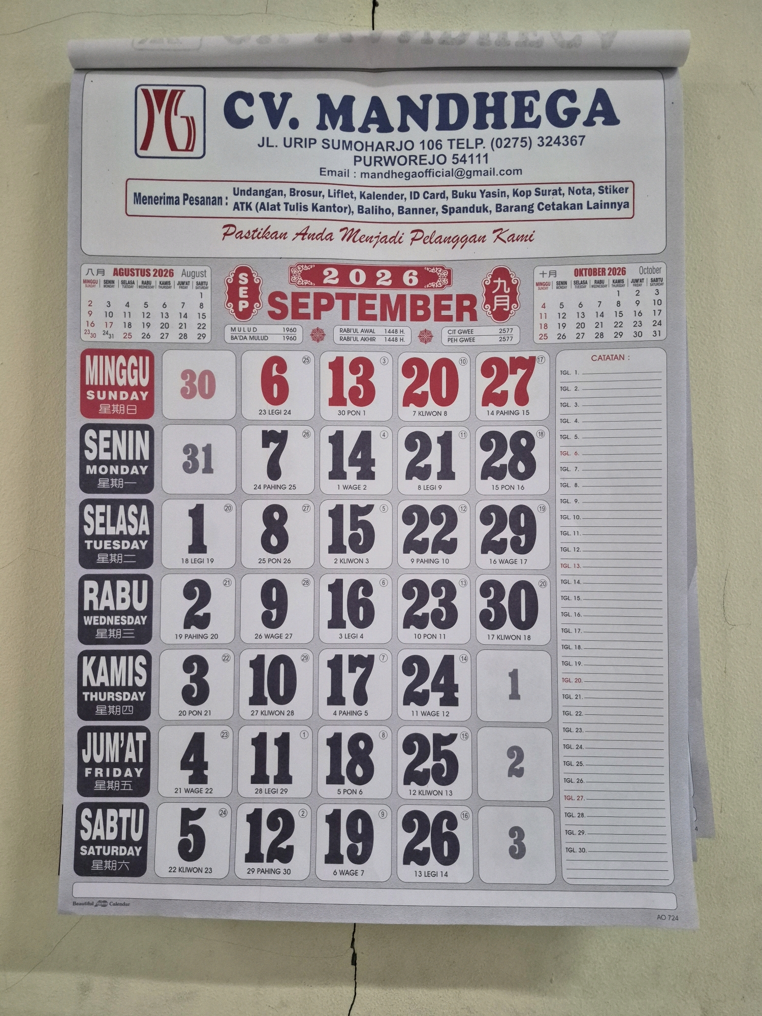 The image displays a wall calendar for the year 2026. The calendar is oriented vertically and features a company logo and name at the top: "CV. MANDHEGA". Below the company name, the address "JL. URIP SUMOHARJO 106 TELP. (0275) 324367 PURWOREJO 54111" indicates the company's location in Purworejo, Indonesia. An email address, "mandhegaofficial@gmail.com", is also present. The calendar prominently features the months of September, October, and November. The September section shows the dates arranged in a grid with days of the week in Indonesian and English. The October section is also visible with its dates and days. The remainder of the calendar shows the beginning of November. A section for "CATATAN" (Notes) with numbered lines is present on the right side of the calendar. The calendar is mounted on a textured, light-colored wall, likely indoors. The overall scene is that of a typical office or business environment displaying promotional material.