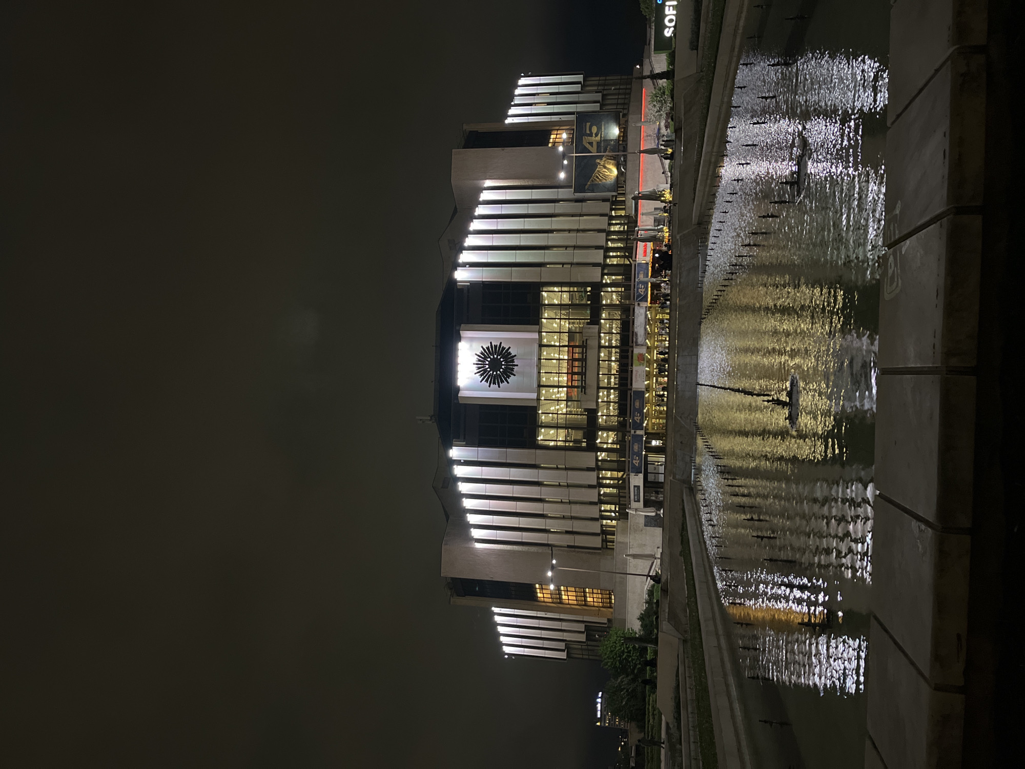 A nighttime photograph captures a modern, illuminated building situated beside a long water feature. The sky is dark, enhancing the bright artificial lights of the structure. The building displays contemporary architecture with large glass sections and prominent vertical white panels that are brightly lit. A distinctive large, circular emblem or decorative element, possibly a clock, featuring radiating spokes, is visible on the lower-middle part of the building's facade.
In the foreground, a rectangular water canal or fountain extends, with numerous small jets creating ripples on its surface. These ripples beautifully reflect the building's lights, forming shimmering patterns across the water. The edges of the water feature are constructed from concrete.
On the upper right, a sign clearly displays the text "SOFIA," indicating the location is Sofia, Bulgaria. Multiple banners or signs on the building's facade prominently feature the number "45." A few indistinct figures can be observed near the building, suggesting general pedestrian presence. The scene evokes a serene and modern urban evening atmosphere, with the illuminated architecture and reflective water creating a strong visual focal point against the night sky.