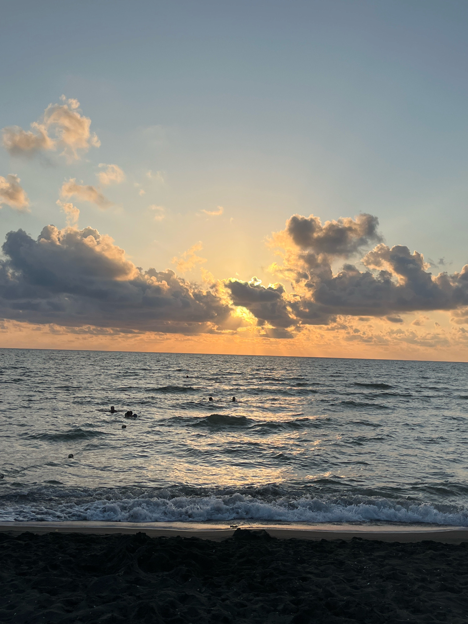 The image depicts a sunset scene at Ureki, Georgia, a Black Sea resort town known for its magnetic sand beaches. The sky is a gradient of pale blue to soft orange, with scattered, fluffy clouds tinged with golden light from the setting sun. The sun itself is partially obscured by clouds, casting rays of light across the horizon. The sea is a deep, rippling blue-gray, with small waves lapping at the dark, sandy shore. Several figures are visible in the water, appearing to be swimming or wading. The foreground consists of the dark, wet sand of the beach, with some texture and footprints visible. The overall atmosphere is serene and tranquil, capturing the beauty of a coastal sunset.