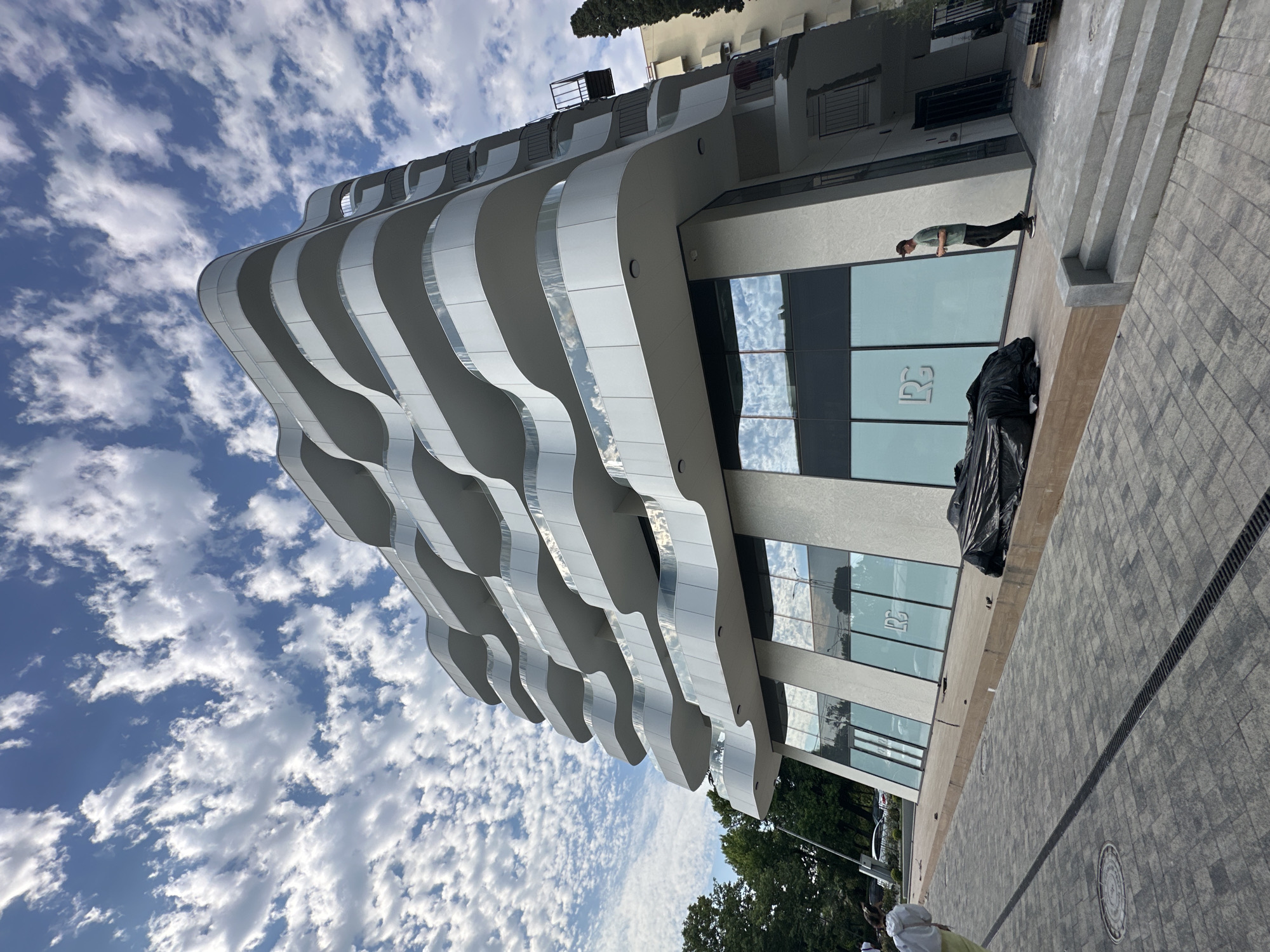 The image depicts a modern, wavy architectural building under a bright, cloudy sky. The building's facade is characterized by undulating white panels that create a dynamic, almost fluid appearance. Large glass windows reflect the sky and surrounding elements.
In the foreground, a person wearing a light green t-shirt and dark pants stands near the building's entrance. They appear to be looking down or towards the ground. A large black tarp or garbage bag is placed on the ground next to the building's windows.
The scene is set on a paved walkway with a patterned stone surface and a drainage grate running along its length. Trees are visible in the background, suggesting an outdoor urban or semi-urban environment. The time of day appears to be daytime, with the bright sky and distinct cloud formations indicating fair weather.
Visible text on the windows includes what appears to be a stylized logo or monogram, possibly "RG". The overall impression is one of contemporary architecture in a public space, possibly during a construction or renovation phase given the tarp. The location is identified as Sochi, Russia.