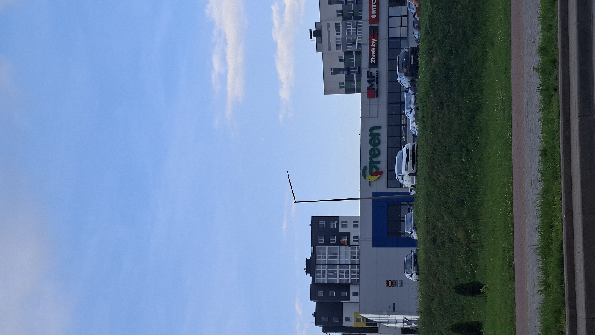 The image shows a street scene in Smalyavichy, Belarus. A modern, multi-story building complex dominates the background, with a large "Green" supermarket visible on the lower level. Several cars are parked in front of the supermarket, and a grassy embankment with trees separates the road from the building. The sky is blue with scattered white clouds. A streetlamp stands in front of the supermarket.