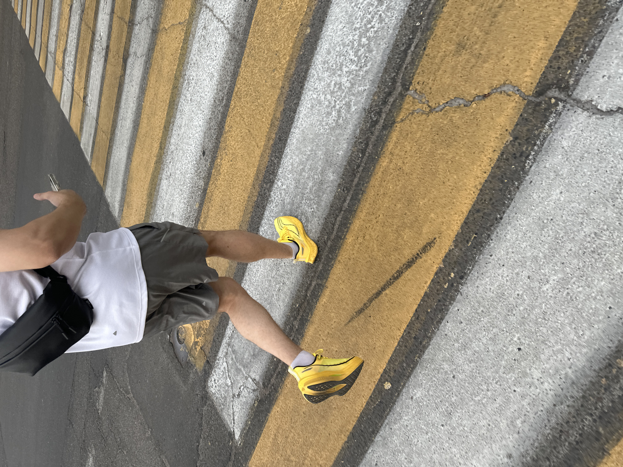 A man wearing yellow athletic shoes, gray shorts, and a white shirt is standing on a pedestrian crosswalk painted with alternating stripes of yellow and light gray. He is holding a cell phone in his left hand and has a black shoulder bag slung across his body. The crosswalk appears to be on a cracked asphalt road. The photo was taken from a high angle, looking down. The location may be Tashkent, Uzbekistan, based on the prompt.