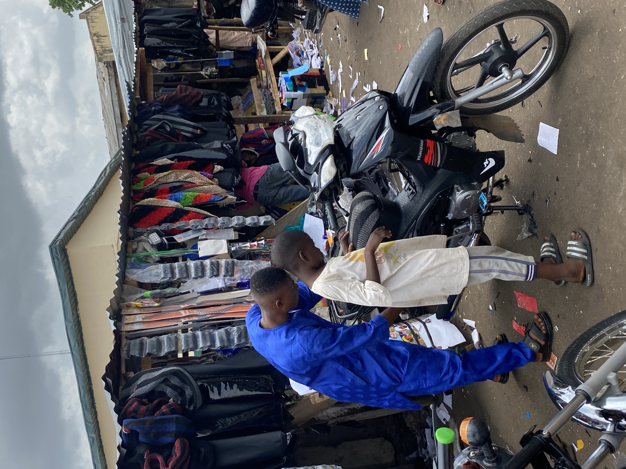 An outdoor market scene in Lokoja, Nigeria, is captured from a slightly elevated, vertical perspective under an overcast sky. The primary subjects are two men or boys standing by a black motorcycle on a dirt-covered ground littered with paper scraps.
The individual on the left wears a striking blue traditional garment, possibly an agbada, and dark sandals. He is positioned leaning towards the motorcycle's handlebars, his hands near the front, appearing to interact with or inspect the vehicle. Next to him, on the right, is another person dressed in a white short-sleeved top and light-colored, striped trousers, sporting black and white Adidas slip-on sandals. This person also faces the motorcycle, with hands resting on its seat.
The prominent motorcycle is black, with "Haojie" visible on its side. To the left, behind the figures, is a market stall with a corrugated metal roof, showcasing a variety of merchandise on wooden shelves and racks. These goods include colorful textile items, seat covers, and other packaged products, indicating a general merchandise or vehicle accessories vendor. A person in a pink shirt is partially visible within the stall. Another motorcycle's front wheel and handlebars are seen on the far right. The ground is dusty and littered, typical of an active open-air market. The time of day is daylight, indicated by the overall brightness, but the sky is cloudy.