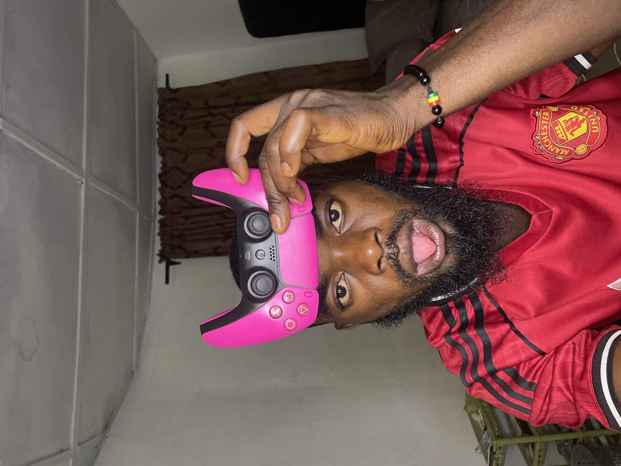 A man of African descent, with dark skin, a full beard, and short black hair, is photographed from the chest up. He is wearing a red Manchester United jersey with black stripes on the sleeves and a white trim on the cuffs. He is holding a pink PlayStation 5 controller to his forehead with his right hand, his thumb extended. His mouth is open, and his tongue is sticking out slightly. He is wearing a black beaded bracelet with red, yellow, and green beads on his right wrist.
The background appears to be an indoor setting, possibly a bedroom, with a textured ceiling and a wall that is painted white or a light color. There are brown curtains visible on the right side of the frame. The lighting is dim, suggesting it is likely evening or night. The overall impression is a casual, playful pose captured indoors.
The image is set in Jos, Nigeria.