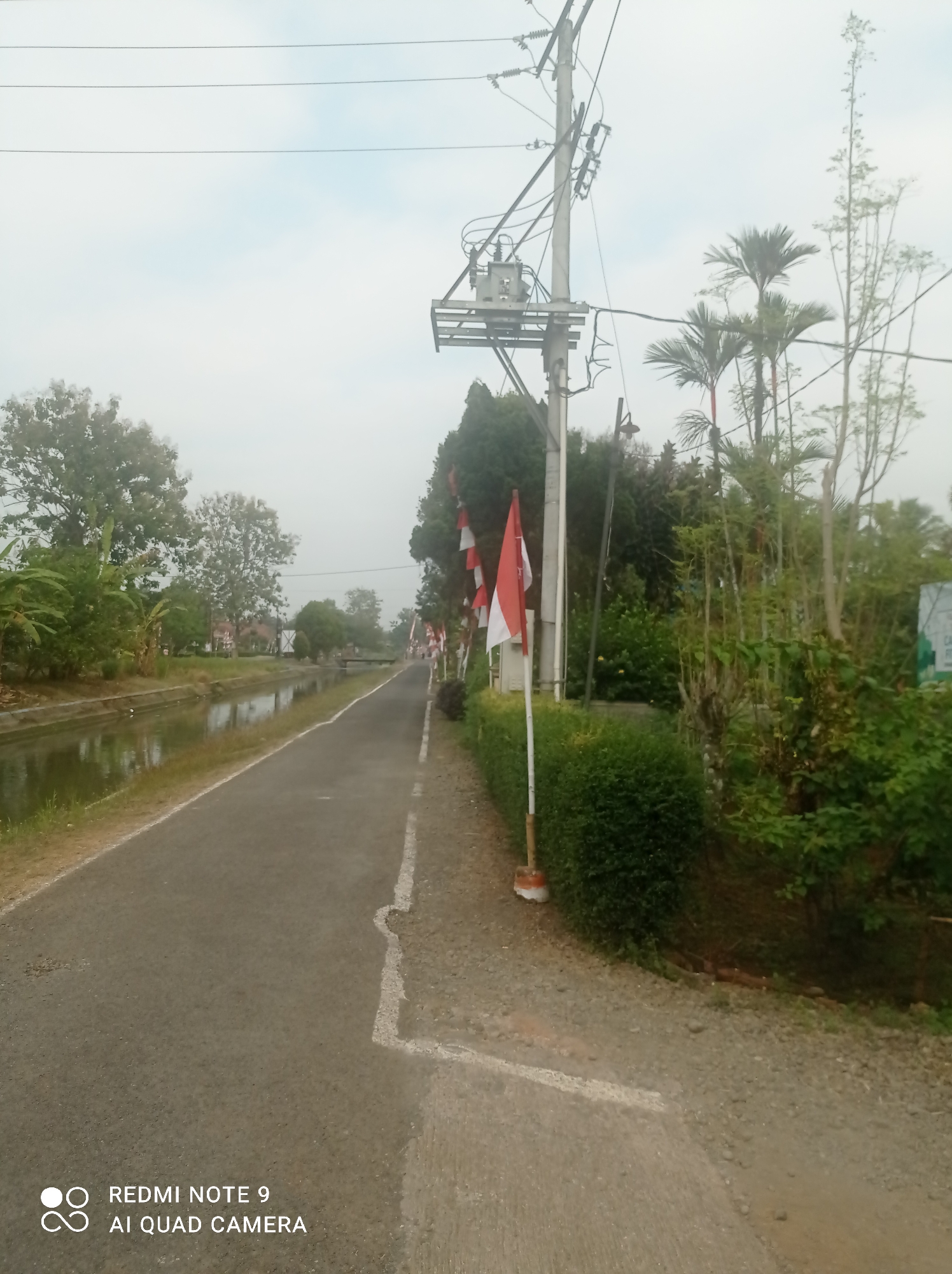 The image depicts a quiet, paved road running alongside a narrow canal. On the left side of the road, lush green trees and vegetation line the canal. On the right side, a neatly trimmed hedge runs along the road, adorned with several Indonesian flags. Beyond the hedge, more trees and vegetation can be seen, along with a utility pole carrying electrical wires and a transformer. The sky is overcast. The scene suggests a peaceful, possibly rural or suburban, setting in Cipawon, Indonesia, likely decorated for a local event or national celebration given the presence of the flags.