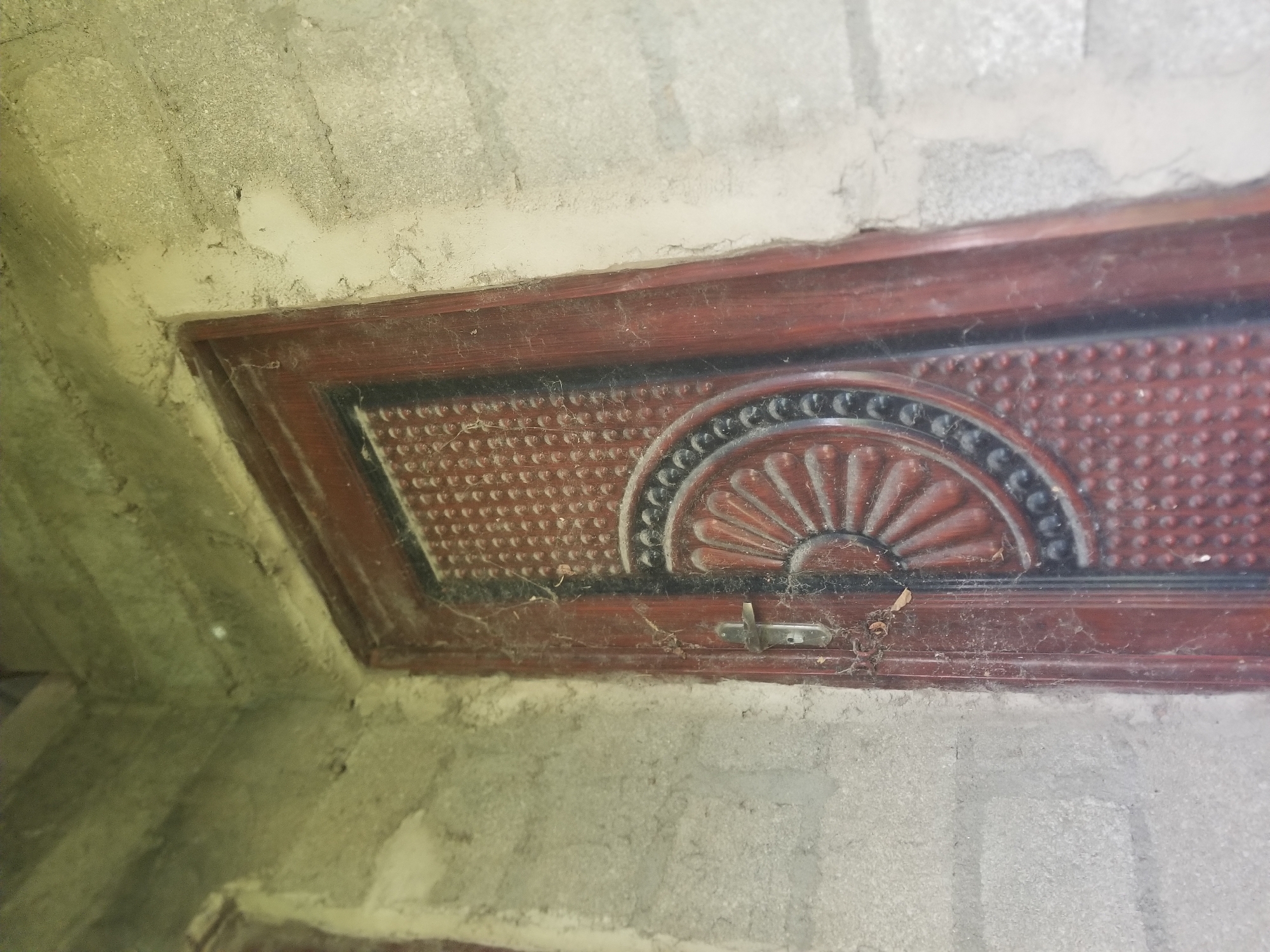 The image shows a close-up view of a dark wooden framed vent or panel set into a rough, textured concrete wall. The vent itself has a decorative, fan-shaped pattern in the center, with a textured, dotted surface filling the rest of the frame. A small, silver latch is visible on the lower edge of the vent. The concrete surrounding the vent appears unfinished, with visible imperfections and variations in texture and color, suggesting an interior or exterior wall in a construction or renovation phase. Cobwebs are present in several areas, particularly around the vent and latch, indicating the area may not be frequently accessed or cleaned. The lighting suggests it is daytime, but the specific time of day or weather conditions are not discernible. There is no visible text in the image. The context of Jalingo, Nigeria, suggests a warm climate, but this is not directly depicted in the image itself. The scene is devoid of any people or overt activity.