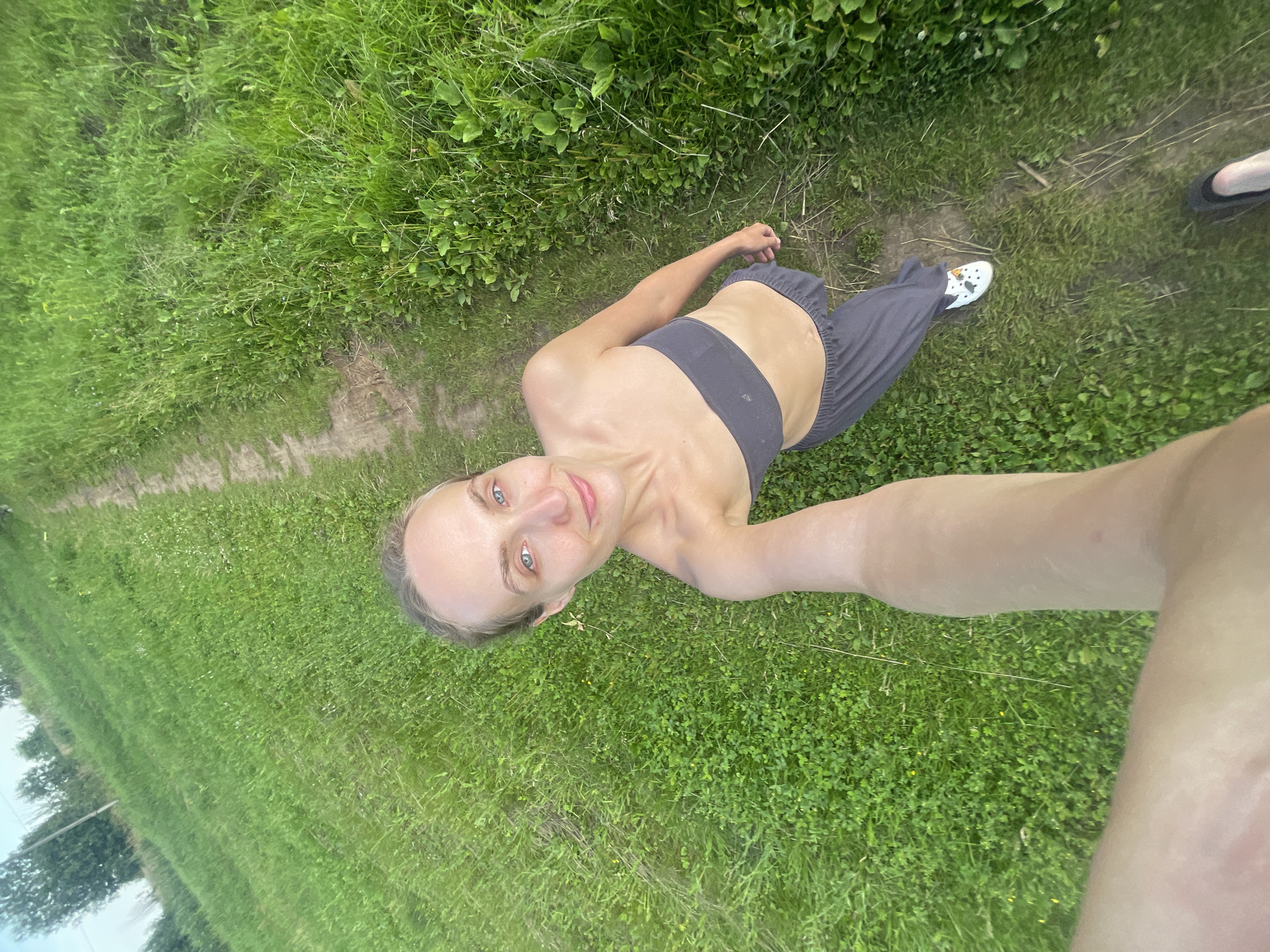 In Подшивалово, Russia, a woman is captured in a high-angle, wide shot, holding a phone to take a selfie. She is standing on a dirt path bordered by lush green grass, with trees and vegetation visible in the background. The woman is wearing a dark gray bandeau top and loose-fitting gray pants. Her hair is pulled back, and she is looking at the camera with a slight smile. She has bright blue eyes and fair skin. Her right foot is visible wearing a white shoe with colorful embellishments, while her left foot is partially obscured by another shoe. The scene suggests an outdoor, possibly rural setting during daytime.
