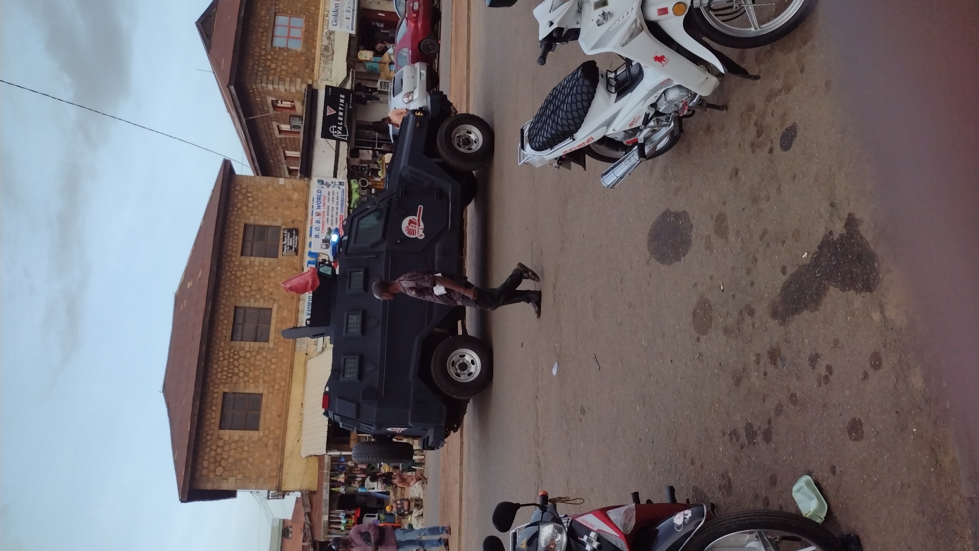 In Jos, Nigeria, a dark, armored police vehicle with flashing blue lights is parked on a paved street. A man in a patterned shirt and dark pants walks past the vehicle, holding a white object. To the left, another person in a maroon shirt and blue jeans stands near market stalls. In the foreground, a white motorcycle is partially visible, and to its left, a red motorcycle is also present. The street is marked with dark oil stains, and a light green disposable container sits on the ground. In the background, two buildings with red tiled roofs are visible, one featuring small windows and the other displaying signage that includes "B.O.B. WORLD CONNECTION LIMITED" and "VALENTINE EMBROIDERY." The sky above is overcast.