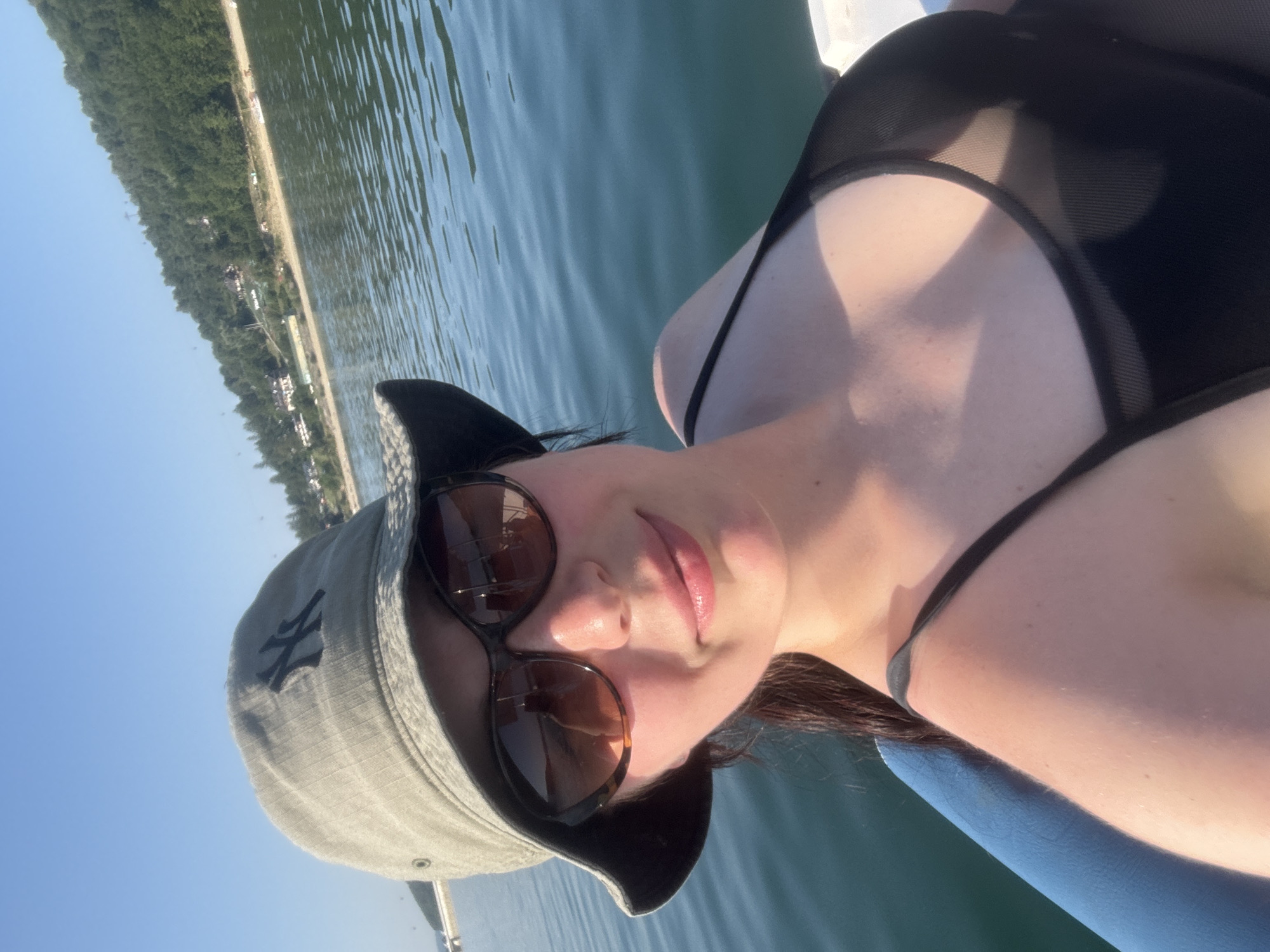 A woman is taking a selfie on a body of water. She is wearing a gray bucket hat with a black New York Yankees logo, sunglasses, and a black swimsuit. She has dark hair and is smiling. The water appears calm, and there is a wooded hill with some buildings in the background. The location is identified as Polańczyk, Poland, a town situated on the Solina Lake reservoir. The sun is shining, casting a warm glow on the woman's face and shoulders. The scene suggests a leisurely day spent outdoors, likely on a boat or a pier.
