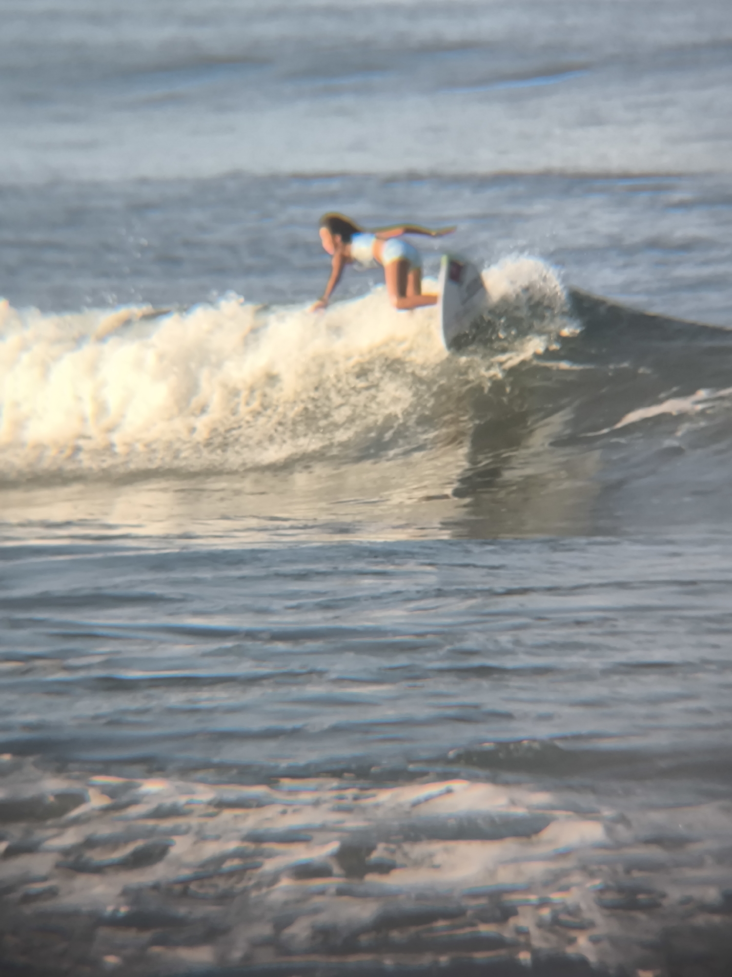A person is surfing on a wave. The surfer, wearing light-colored swimwear, is positioned on a surfboard on the crest of a wave. The wave is breaking, with white water visible. The scene is North Kuta, Indonesia. The ocean's surface is textured with ripples.