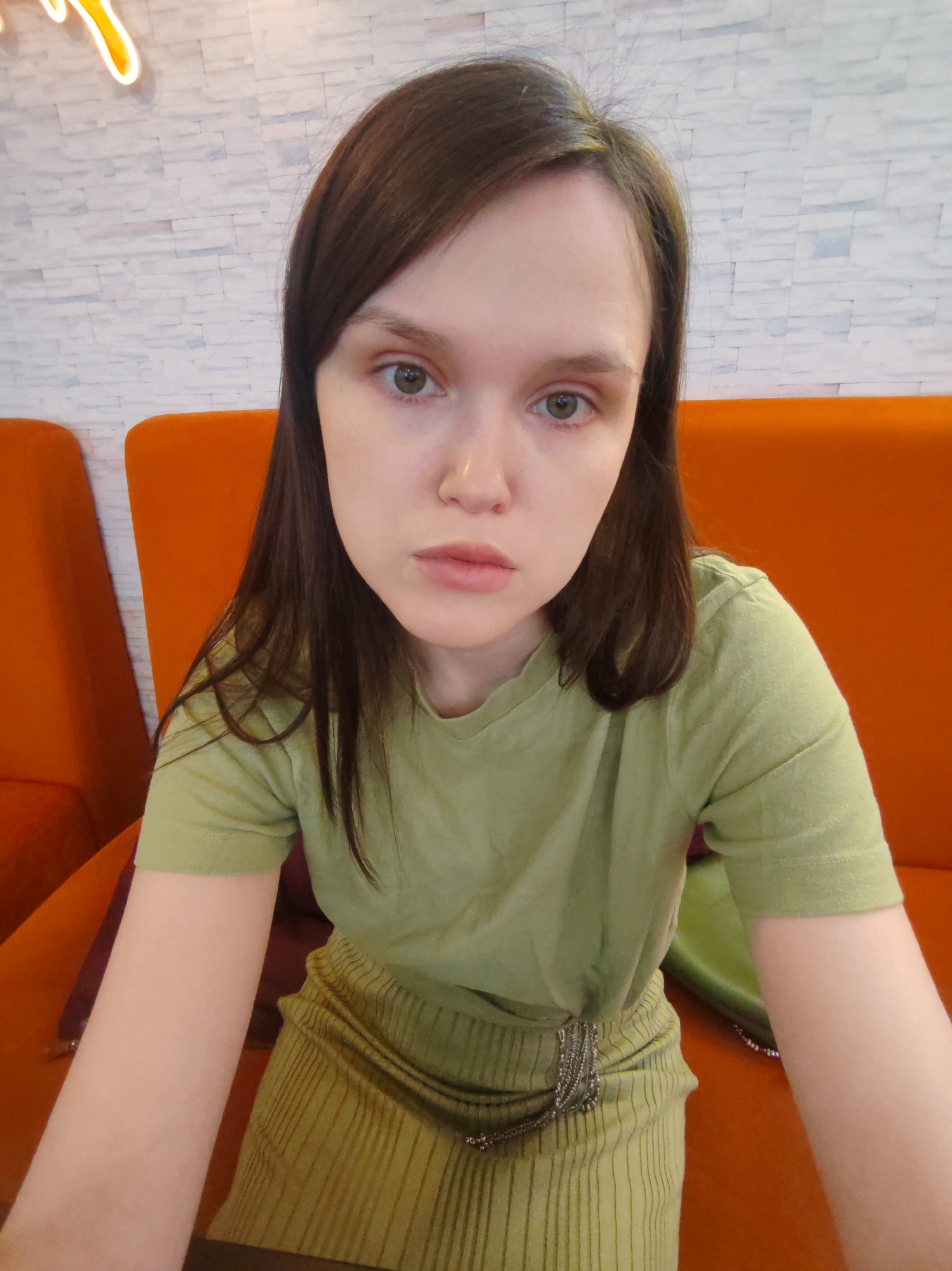A young woman with long, dark brown hair, light skin, and green eyes is centered in a close-up indoor shot, looking directly at the camera with a neutral expression. She is wearing a light green short-sleeved top and a matching ribbed skirt or dress, cinched at the waist with a silver-colored chain or decorative belt.
She is seated in a booth or on a couch, which, along with another identical piece of furniture partially visible to the left, is upholstered in a vibrant, bright orange fabric. Behind her, a light-colored, textured wall, resembling white faux brick or stone, serves as the background. In the upper left corner, a portion of a glowing orange neon sign with an abstract, curved design is visible. The overall setting appears to be an indoor commercial space, possibly a cafe or restaurant in Ust-Ilimsk, Russia, characterized by its modern and casual decor. No discernible text is visible in the image.