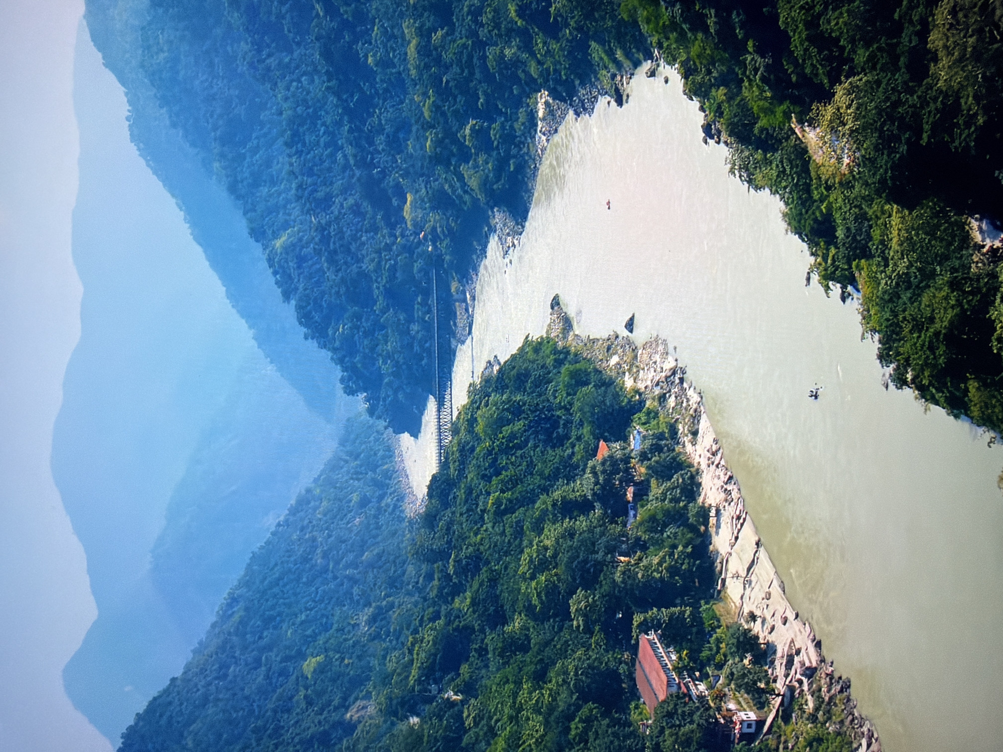 The image depicts a wide, meandering river flowing through a lush, mountainous valley. The river's water is a light, sandy color, and its surface is relatively calm with a few small ripples. Along the left bank, there are several buildings with red roofs nestled amongst the trees. A rocky shoreline with some patches of sand borders the river. In the distance, a suspension bridge spans the river.
The valley walls are densely covered in green foliage, with trees and undergrowth creating a thick carpet. The mountains in the background are layered, fading into a hazy blue at the horizon, suggesting a significant distance and atmospheric perspective. There are a few small boats visible on the river, and what appears to be a single person on a raft further downriver.
This image does not depict any location in the city of Kyiv, Ukraine. The landscape is characteristic of a mountainous river valley, possibly in Asia, given the architecture of the buildings.