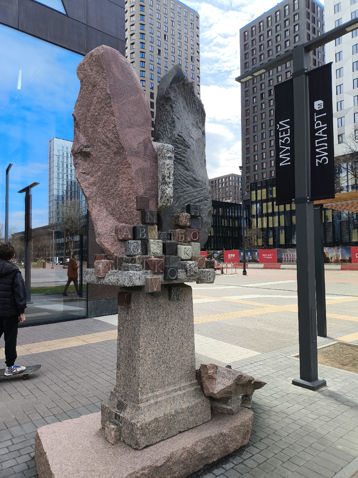 A full-length outdoor shot reveals a city square in Moscow, Russia, adorned with a stone sculpture and urban structures. A boy in a black puffer vest and pants stands on a skateboard, gazing at the sculpture. The sculpture features two large, rough-hewn stones, one pinkish-brown and the other gray, stacked on a stone base. Between them, several stone blocks with Cyrillic letters form a horizontal layer.
In the background, a modern glass building reflects the sky, and a woman in a brown coat walks by. High-rise buildings populate the cityscape. To the right, a dark metal pole holds two vertical black banners that say "МУЗЕЙ" and "ЗИЛАРТ" (Museum and ZILART), indicating the proximity to the ZILART area in Moscow. The ground is paved with gray and tan rectangular stones.
