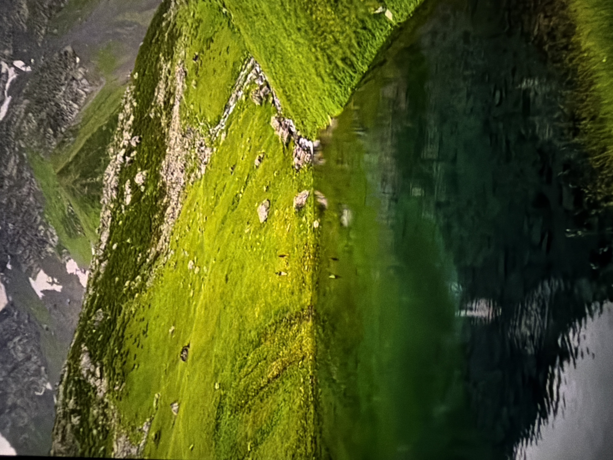 The image shows a landscape with a green, grassy hillside next to a body of water, likely a lake. The hillside is dotted with rocks and possibly a small stream or path. In the background, there are mountains with patches of snow. The water reflects the hillside and sky, creating a sense of depth and tranquility. The location is not specified.